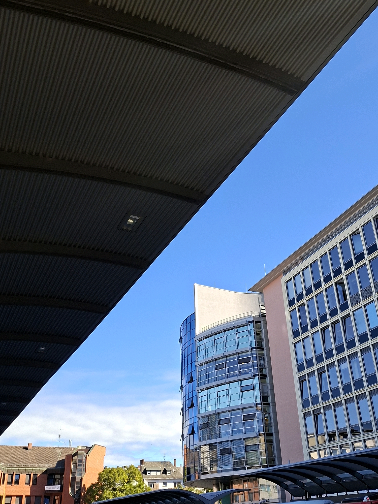 The image depicts an exterior view looking upwards towards the sky and buildings. In the foreground, a dark, ribbed metal structure, likely a roof or overhang, dominates the left side of the frame, casting a shadow. A light fixture is visible on this structure. To the right of this structure, a clear blue sky with a few wispy clouds is visible.
In the mid-ground, there are two modern office buildings. The building on the left is characterized by its curved glass facade, reflecting the sky and surrounding structures. It has multiple floors with visible windows, some of which are open. Adjacent to it on the right is another office building with a more rectilinear design, featuring a repetitive pattern of windows with dark frames. This building has a lighter colored exterior wall with a pinkish hue on one section.
Further in the background, a cluster of older brick buildings with tiled roofs is visible. These buildings appear to be lower in height than the modern office buildings. A vibrant green tree can be seen amongst these buildings, adding a touch of nature to the urban landscape.
The image is taken on a bright, sunny day. Based on the architectural style and the presence of the "TALGO" signage on one of the brick buildings in the background, the location is identified as Koblenz, Germany, specifically the area near the Koblenz Hauptbahnhof (main train station), likely a bus or tram stop. There are no people visible in the frame. The scene is a typical urban environment with a mix of old and new architecture.