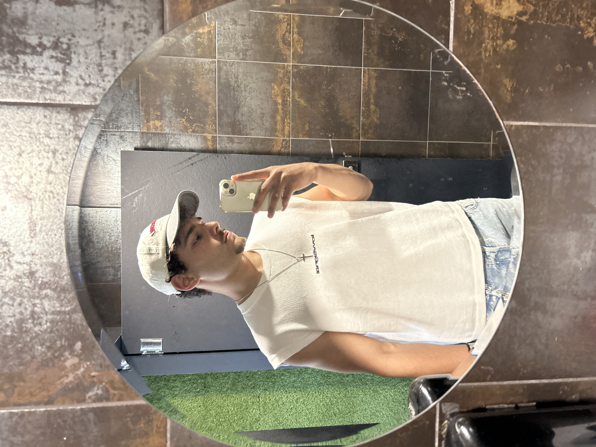 A young man is captured in a mirror selfie within a round mirror. The image, originally rotated, depicts him standing in an indoor setting. He appears to be in his late teens or early twenties, with dark hair and a light mustache. He is wearing a white baseball cap with a red logo (partially visible, possibly an 'A'), a plain white short-sleeved t-shirt, and light blue denim jeans. A silver chain with a cross pendant hangs around his neck. He holds a light-colored iPhone, identifiable by the visible Apple logo and multiple cameras, to take the photograph.
The reflection in the mirror reveals the surrounding environment. Behind the man, there is a dark, possibly black, wall or partition. Below this, a patch of artificial green grass (turf) is visible. Above his head, the walls are adorned with dark, rectangular tiles that have a textured, somewhat metallic or distressed appearance, showing shades of brown, gold, and black with prominent grout lines. A dark, indistinct object, possibly a counter or sink, is seen in the bottom right of the mirror's reflection.
The man's expression is neutral and calm, as he gazes slightly upwards and to the side, not directly at the phone's camera. The setting suggests a modern, well-lit indoor facility, such as a gym, sports complex, or a contemporary commercial space. The location context provided is Sofia, Bulgaria.
Visible text includes the Apple logo on the phone and a word on the t-shirt that, due to the mirror's reflection, appears as "anplewol" or a similar reversed spelling. A red symbol on the cap is also visible.