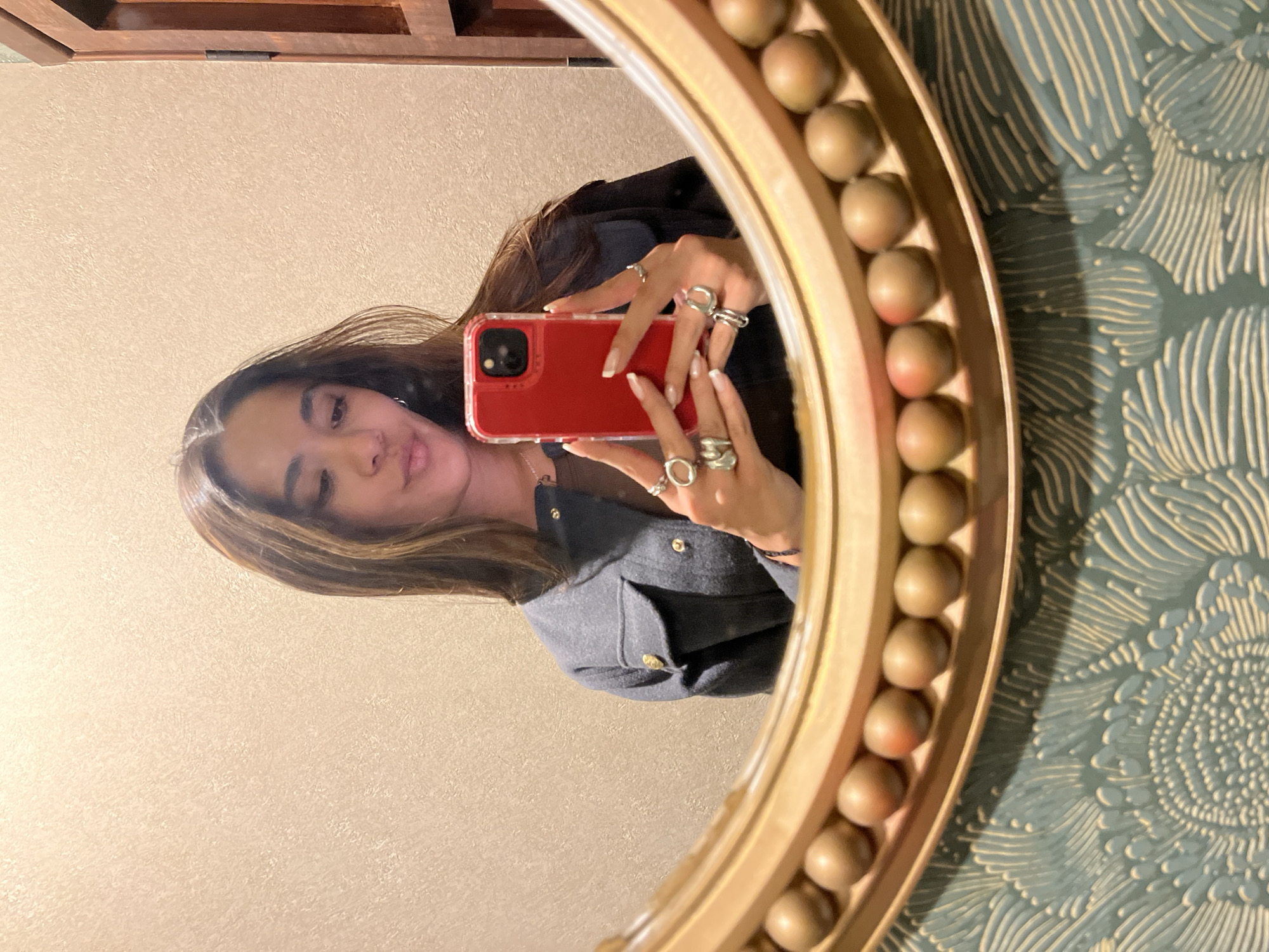 A woman is taking a selfie in a round, gold-colored mirror with a decorative border consisting of round balls. She is holding a red smartphone in her hands, which are adorned with multiple rings. The background behind the mirror shows a patterned blue and white wallpaper on the right side, and a plain beige wall on the left side. A portion of a brown wooden cabinet is visible at the top of the frame. The woman has brown hair with blonde highlights. She is wearing a gray jacket. The location cannot be determined solely based on the image.