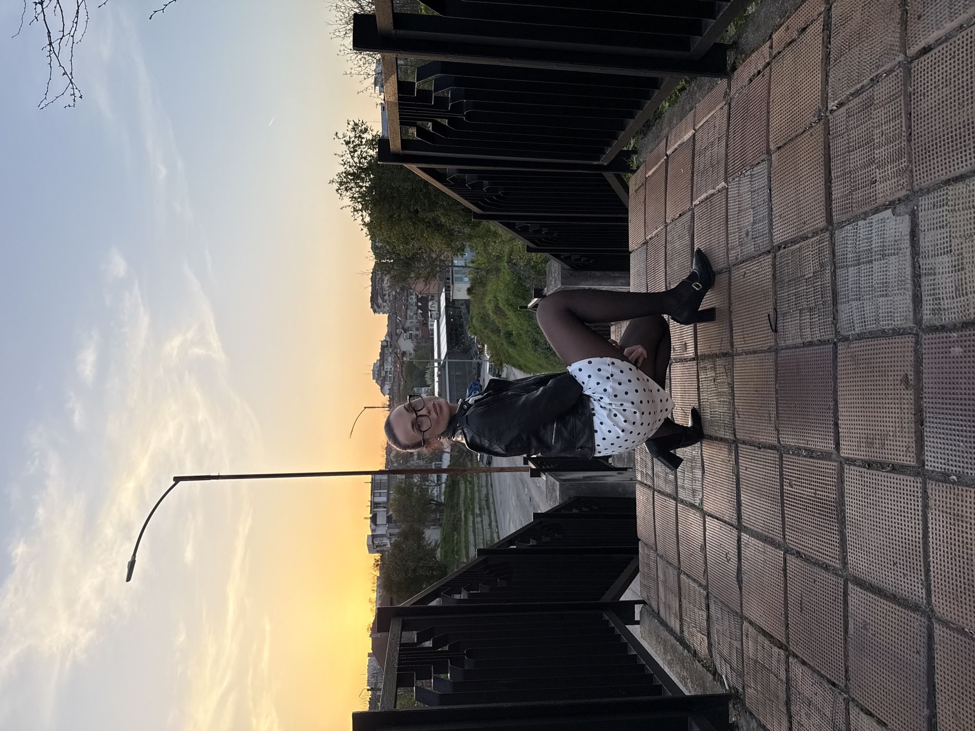 A captivating full-body portrait captures a young woman seated casually on a tiled ledge in Pleven, Bulgaria, under a picturesque sky. She is positioned with her legs bent to her left, looking directly at the camera with a calm expression, framed by glasses. Her attire includes a black leather jacket, a white dress or skirt adorned with black polka dots, black tights, and black high-heeled boots with a buckle detail.
The woman is situated beside a robust dark metal railing or fence, which separates her from a sprawling cityscape visible in the midground, featuring various buildings and patches of green foliage. To her left, a tall, dark street light pole with a curved arm extends upwards, silhouetted against the sky.
The upper half of the image showcases a stunning twilight sky, transitioning smoothly from a soft blue with subtle cloud formations on the left to a vibrant orange and yellow glow towards the horizon on the right, indicating either sunrise or sunset. The light casts a warm, serene ambiance over the entire scene. The foreground features textured, rectangular paving stones with a grid pattern. No visible text is present in the image.