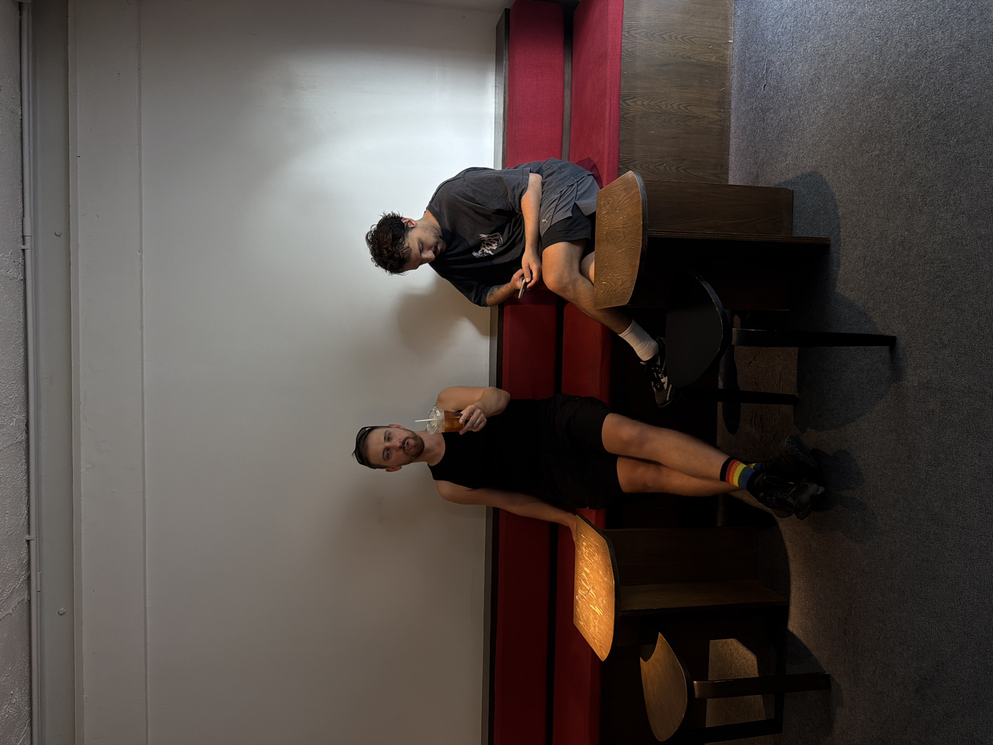 Two men are seated on a red bench in a cafe. The man on the left, wearing a black tank top and black shorts, holds a clear plastic cup with a straw. He is also wearing rainbow-striped socks and black sneakers. The man on the right, wearing a dark grey t-shirt and dark grey shorts, is looking down at his phone. Both men are sitting at separate wooden tables. The cafe appears to have a minimalist design with white walls and wooden accents. This scene is located in Chiang Mai City Municipality, Thailand.