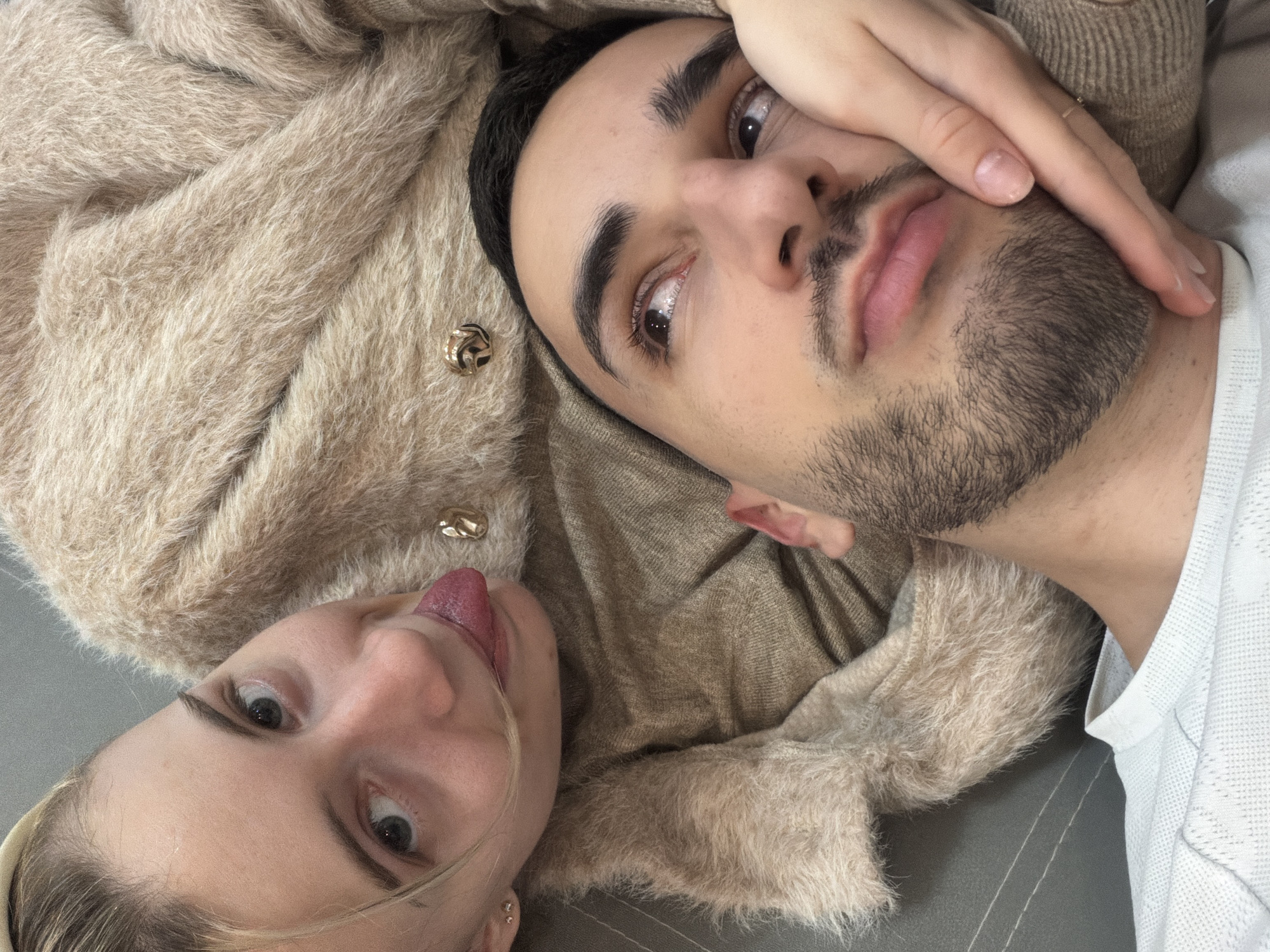 A selfie-style photograph shows a woman and a man lying down. The woman is on the left, with her head tilted towards the camera and her tongue sticking out. She has fair skin, light-colored hair, and noticeable eyebrows. The man is on the right, with his hand partially covering his face. He has dark hair, dark eyebrows, and a short beard. Both are looking at the camera. They are lying on a surface that has subtle horizontal lines. The woman appears to be wearing a furry, light brown garment, while the man is wearing a white top. The location cannot be determined from this image.