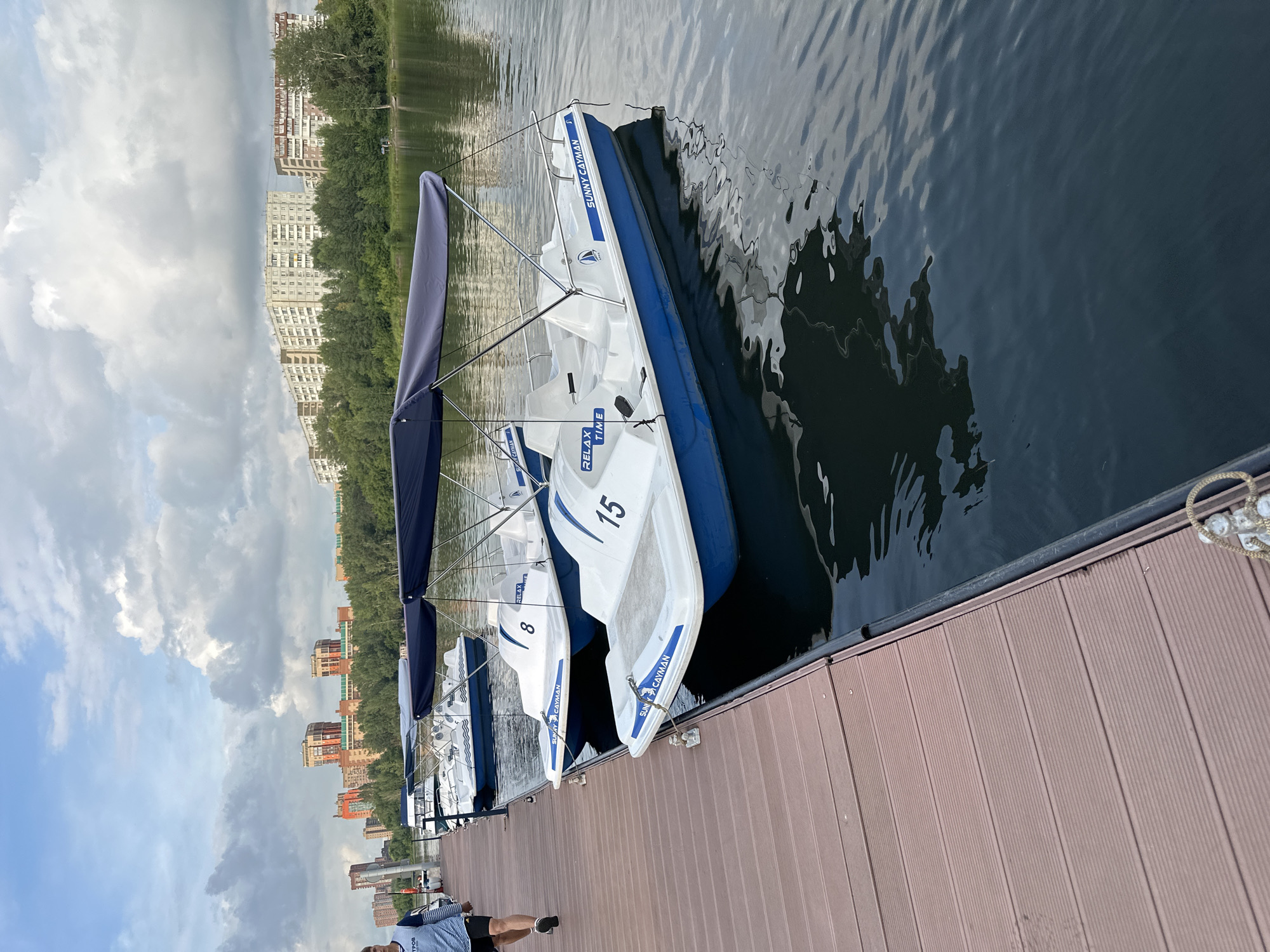 A person walks along a wooden pier next to a row of parked, empty pedal boats on a body of water. The pedal boats are white with blue trim and have navy blue canopies. Several buildings and trees are visible in the background across the water. The sky is cloudy.
