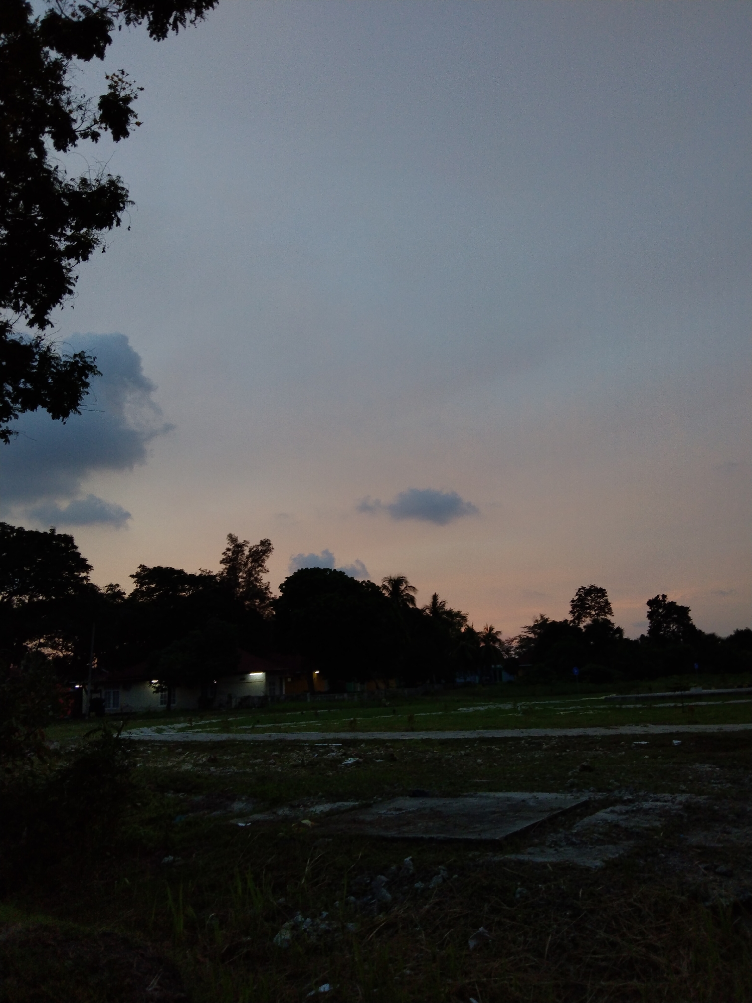The image captures a tranquil dusk or early evening scene, likely in a rural or semi-urban area of Lhokseumawe, Indonesia. The sky dominates the upper two-thirds of the frame, displaying a gradient of colors from a deep, muted blue-grey at the zenith to soft orange and pink hues near the horizon, characteristic of twilight. Scattered dark grey clouds are visible against the fading light.
A thick line of silhouetted trees forms the mid-ground, their dark forms contrasting sharply with the sky. A large tree branch with leaves extends into the frame from the upper left corner, also in silhouette. Nestled among these trees are several low-rise buildings, appearing to be houses or small structures. Artificial lights are visible from within or on the exteriors of these buildings, casting a warm, yellow glow that stands out in the diminishing natural light, further indicating the late hour.
The foreground consists of an open, unkempt field or vacant lot. Patches of dark grass and bare earth cover the ground, with some concrete slabs or structures visible, possibly remnants of construction or foundations. There is no visible activity or people in the scene, contributing to a sense of peaceful stillness. The overall atmosphere is quiet and serene as day transitions to night. No legible text is visible in the image.