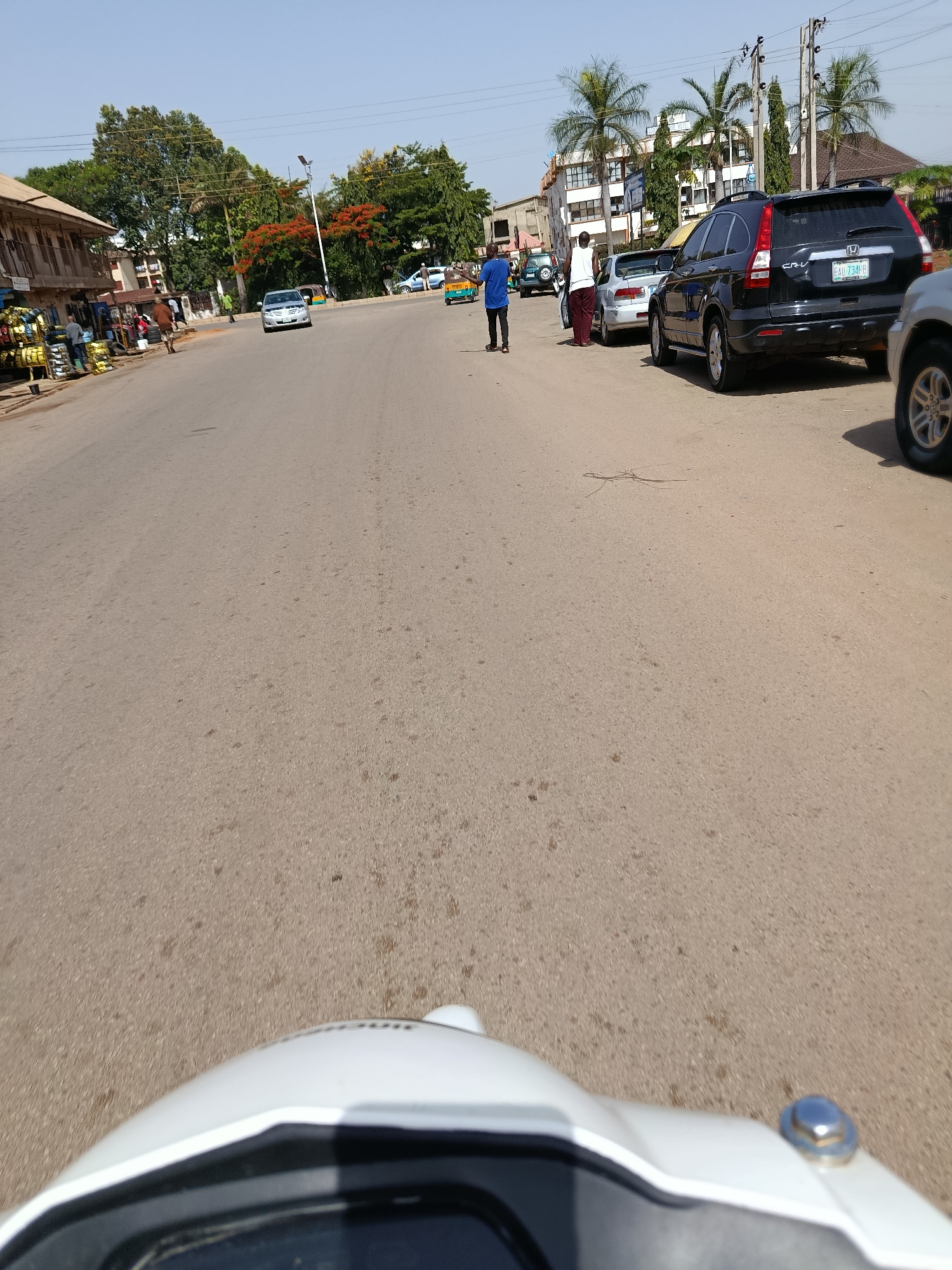 In the city of New Stadium, Nigeria, a street scene unfolds under a clear sky. The perspective is low, suggesting it was taken from a motorcycle or similar vehicle, with the front of a white vehicle dominating the foreground. The paved road is the primary focus, showing signs of wear and scattered debris.
To the left, a row of single-story buildings with open fronts lines the street, displaying various goods for sale, including tires and other items. Several individuals are visible, some interacting with the stalls, others walking along the road. A light-colored car is parked on the side of the road.
Further down the street, a silver car is moving, and behind it, a black SUV, identified by its license plate "EAU-734 JB", is parked. Another person in a white shirt and dark pants stands near the black SUV. A person in a blue shirt is walking away from the camera, near a yellow and blue rickshaw. Palm trees and lush green trees with some red blossoms are scattered throughout the background, interspersed with buildings, some of which appear to be multi-story structures. Utility poles and overhead wires are also visible. The overall impression is of a busy, everyday street in a Nigerian city.