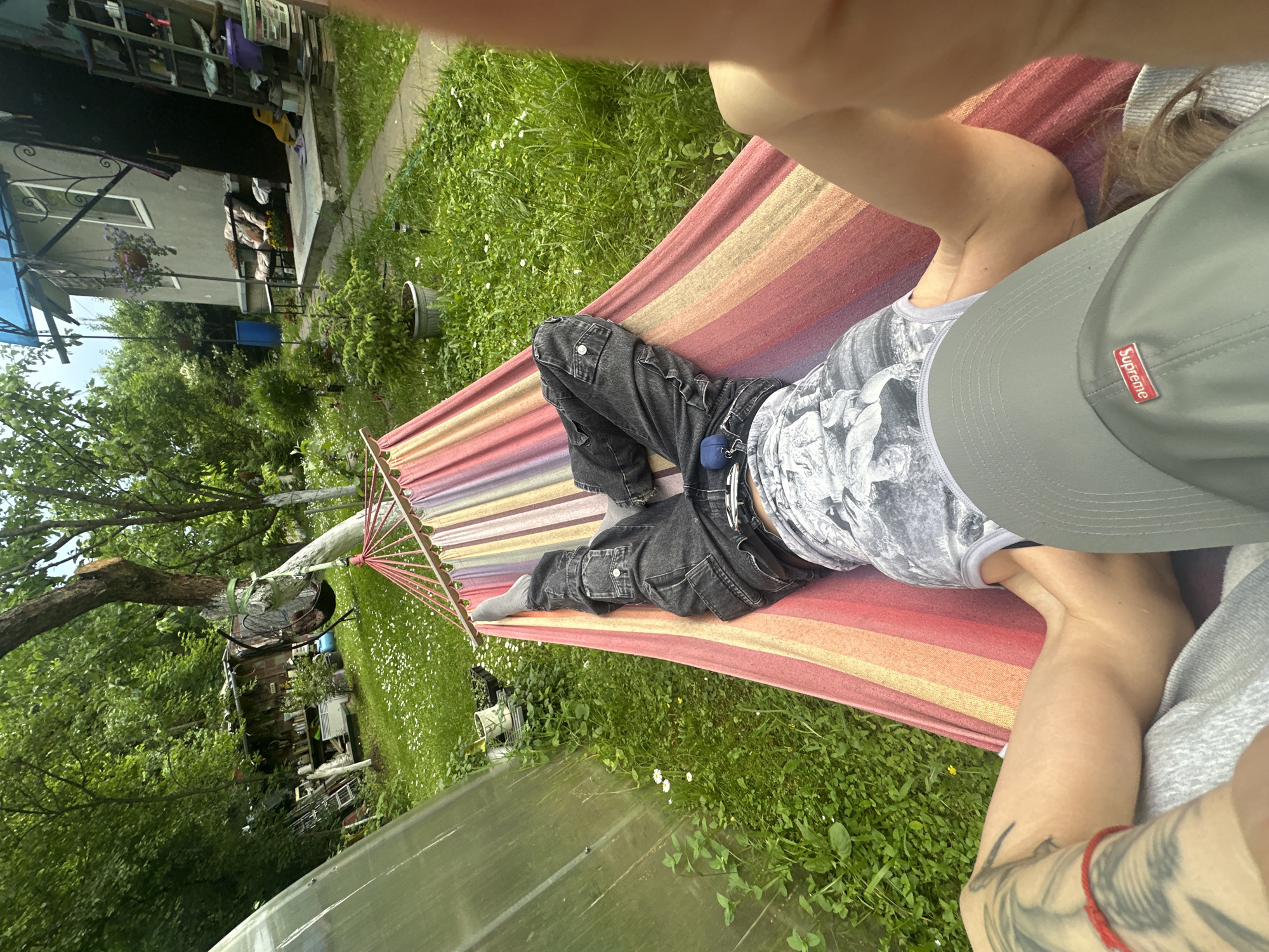 In Козино, Russia, a person is lying in a colorful hammock in a backyard. The hammock is suspended between two supports, with a tree branch visible on the left and what appears to be another support on the right, partially obscured by the photographer's arm. The person is wearing dark jeans, grey socks, and a grey baseball cap with a red "Supreme" logo. Their legs are crossed, and they are holding a blue object with their left hand. The background shows a grassy area with scattered white flowers, a greenhouse structure to the left, and a building with a porch and various items stored outside to the right. Trees and lush green foliage surround the scene, indicating a natural, outdoor setting. The image is taken from a first-person perspective, looking down at the person in the hammock.