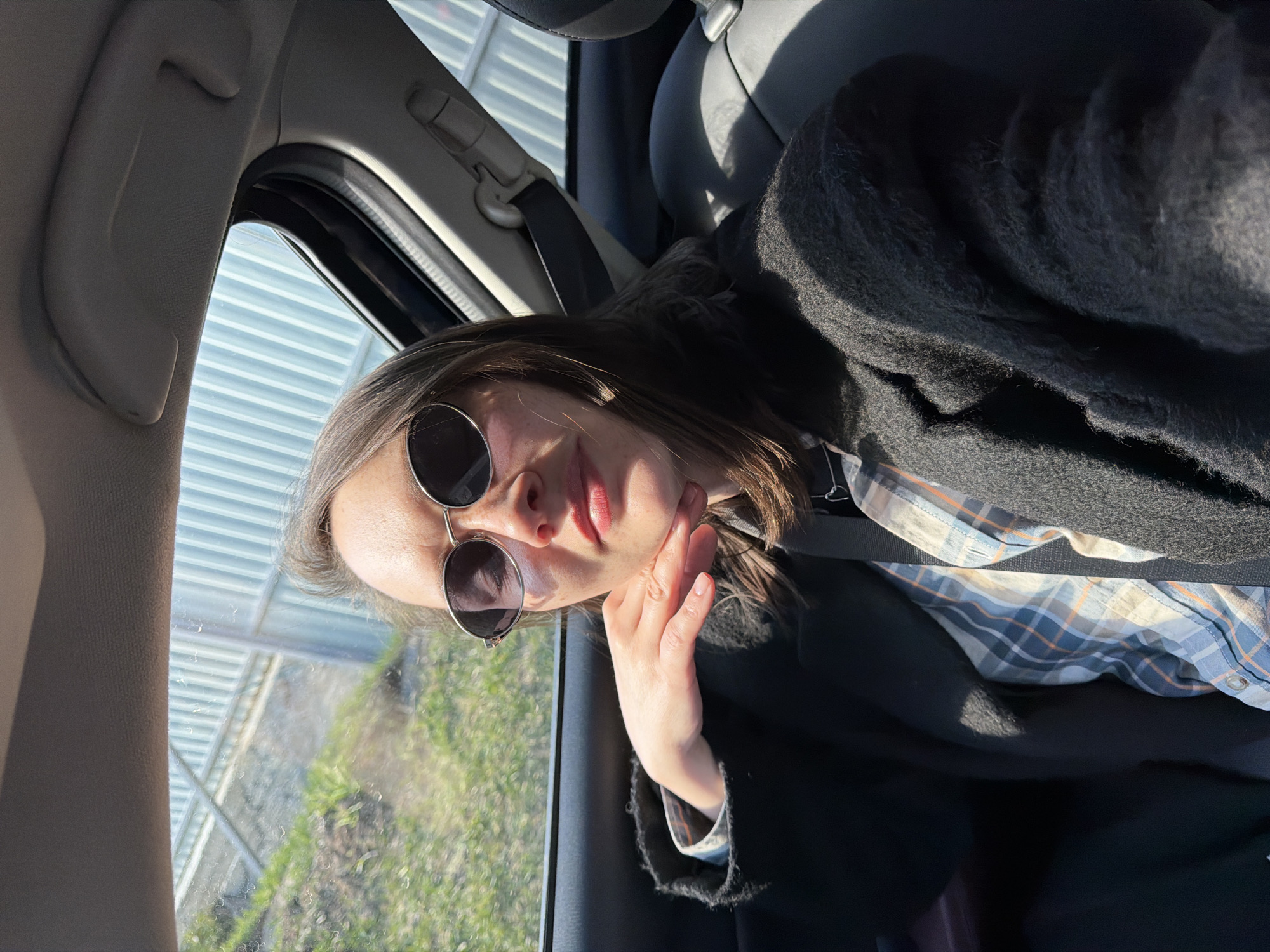A woman is taking a selfie from inside a car. She is wearing sunglasses, a plaid shirt, and a black coat. The window is visible, showing a view of a building and some greenery outside. The location is likely in Chaikovsky, Russia, as specified in the prompt.