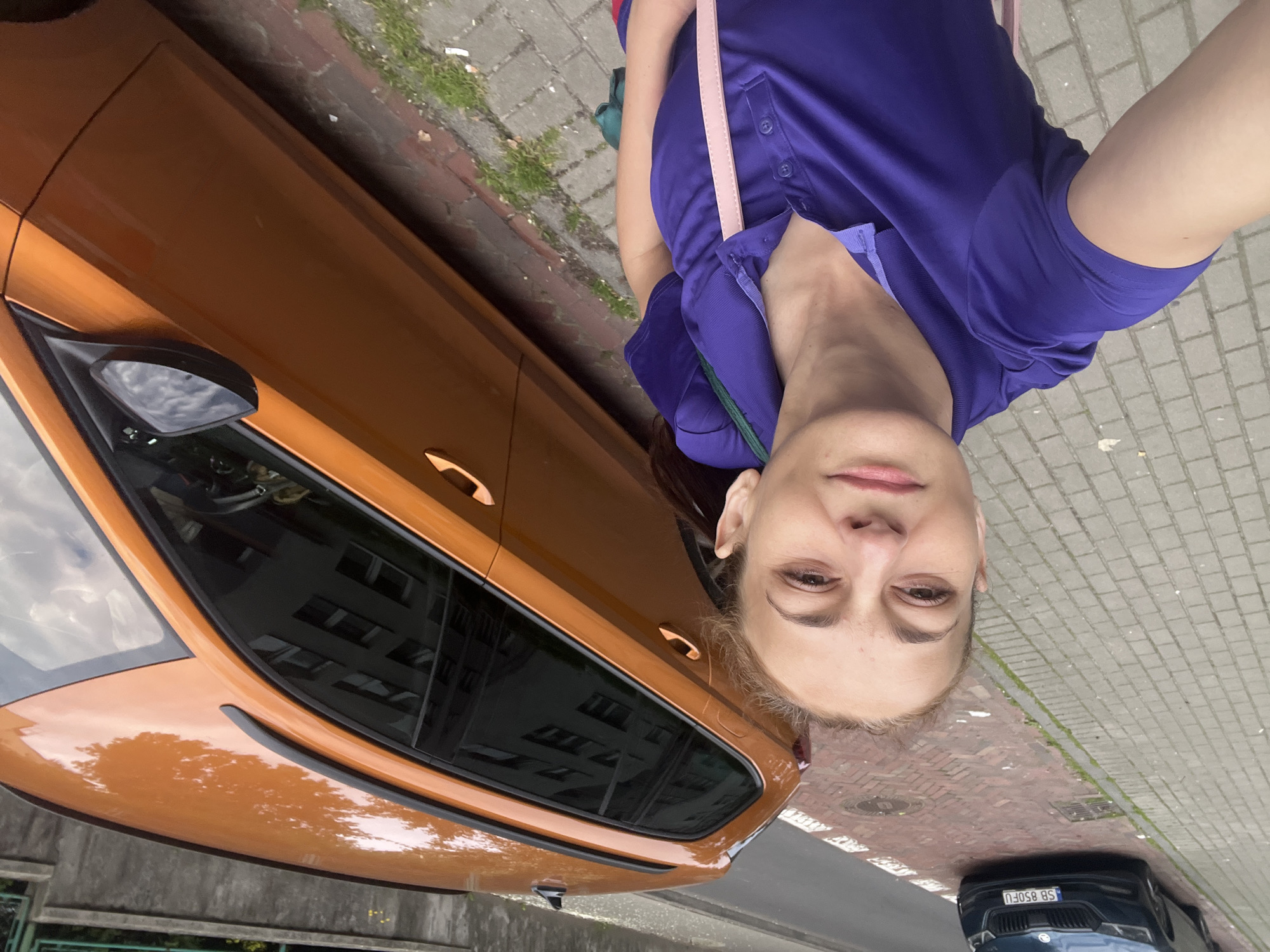 A woman wearing a purple polo shirt stands on a cobblestone sidewalk next to an orange station wagon. The woman is holding a phone in her right hand, taking a selfie. The car's doors and windows are visible. In the background, another dark-colored car is parked on the street, with the license plate "SB 850FU". The environment appears to be an urban setting with a building visible in the reflection of the orange car's windows. The sidewalk has some grass growing between the cobblestones.