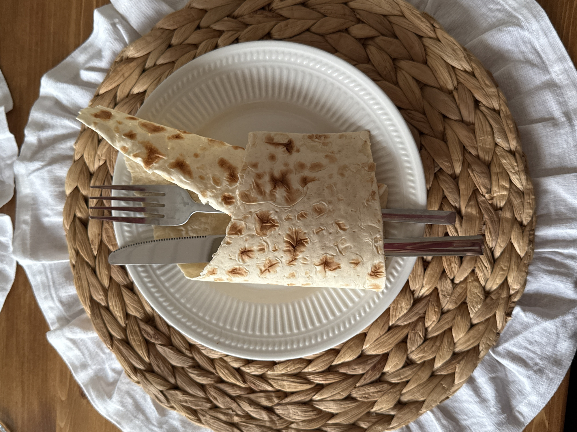 A top-down, eye-level shot captures a meal setting on a wooden surface. A woven, round placemat made of straw-colored material sits on top of a white linen napkin. Centered on the placemat is a white, fluted plate.
Resting on the plate are two pieces of flatbread, which appear to be lavash or a similar type of unleavened bread. One piece is folded diagonally, while the other is folded into a rough square shape. Both pieces have visible char marks from being cooked. A fork and a knife are placed on top of the flatbread, with the fork on the left and the knife on the right, suggesting the meal is ready to be eaten. The flatbread is the primary subject, with the plate, cutlery, placemat, and napkin serving as supporting elements that create a sense of a prepared meal.
The lighting suggests it could be daytime, with diffused light casting soft shadows. The overall impression is one of a simple, rustic meal presentation. There is no visible text in the image. The location context is Starokorsunskaya, Russia.