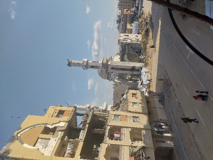 The image depicts a scene of extensive urban destruction in Khan Yunis, Palestinian Territory, under a clear blue sky with scattered white clouds.
In the left foreground, a multi-story building, light yellow-beige in color, is severely damaged, with large sections of its facade and interior floors collapsed and exposed. Laundry hangs from balconies on multiple levels. An arched architectural detail is visible on its upper section. The ground level features a mural with red, white, and blue geometric shapes, and a bicycle is parked near the entrance. Piles of concrete and other debris are scattered along the sidewalk and street edge beside this building.
Across a paved street with white lane markings, the central feature is a severely damaged mosque. Its light-colored minaret stands partially intact but shows structural damage, while the main body of the mosque, including its dome, is largely reduced to rubble. Extensive piles of debris surround the mosque's remains. Amidst this rubble, several makeshift shelters, appearing to be constructed from tarpaulins over frames, are visible.
On the paved street, an adult male in dark clothing walks away from the viewer on the left side of the road. A group of four children stands together in the middle of the street; three wear red tops, and one wears a blue top. Another individual walks through the dusty, rubble-strewn area to the right of the mosque. In the background, further damaged buildings and urban structures stretch into the distance.