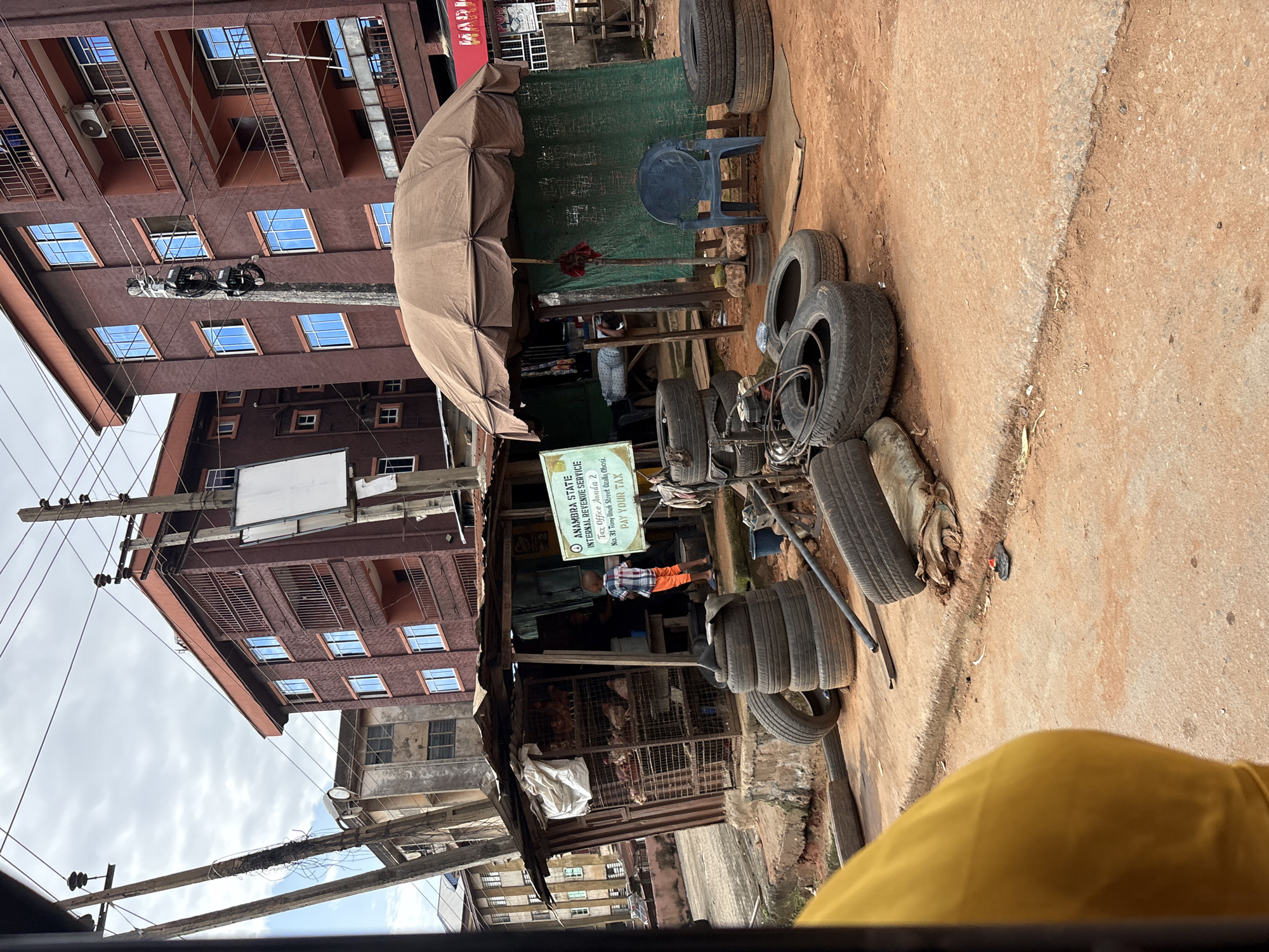 The image depicts a street scene in Nkpor, Nigeria, with a focus on a small roadside stall and its surroundings. The stall appears to be selling poultry, with chickens visible in cages. In front of the stall, a stack of tires is piled up, suggesting a tire repair or sales business operating alongside the poultry. A young boy in a checkered shirt and orange shorts stands near the stall. To the right of the stall, a blue plastic chair is placed under a brown canopy, and a green fabric serves as a backdrop. Electrical poles and wires crisscross the sky above. The ground is unpaved and reddish-brown. In the background, multi-story buildings with reddish-brown facades and blue-tinted windows are visible. A sign in the foreground reads "ANAMBRA STATE INTERNAL REVENUE SERVICE" and "PAY YOUR TAX". The overall impression is of a bustling, informal commercial area.