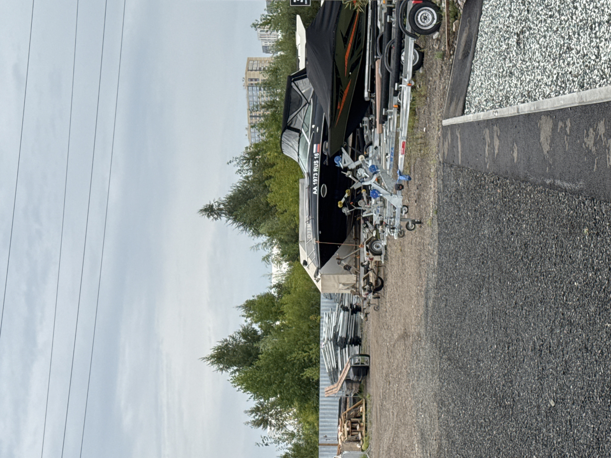A boat storage lot is pictured in Izhevsk, Russia, under an overcast sky. The lot is filled with boats on trailers and stacks of what appear to be boat parts or construction materials. In the foreground, dark grey gravel covers the ground, transitioning to asphalt and then light grey gravel near a concrete barrier. Several boat trailers with wheels are visible. One boat, predominantly black with orange accents, is covered with a black tarp. Another boat, also black, is partially covered and displays the text "AA 1973 RUS 16" on its side. In the mid-ground, stacks of metallic materials and wooden pallets are arranged near a corrugated metal fence. Beyond the fence, a dense line of green trees forms a backdrop. In the distance, tall residential buildings with light-colored facades are visible, suggesting an urban or suburban setting. Overhead, four parallel power lines stretch across the sky. No people are present in the image.