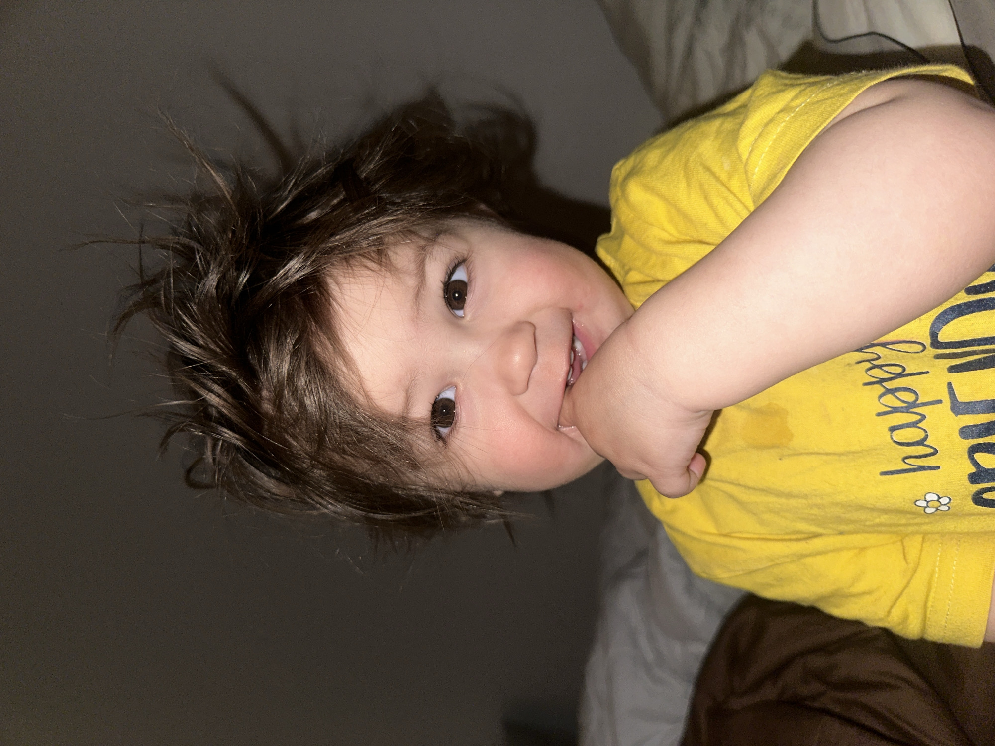 A toddler with brown hair and brown eyes is lying on a bed with bedding in varying shades of brown and grey. The toddler is wearing a yellow t-shirt with writing on it. The toddler is holding their left hand to their mouth. The background is dark and out of focus. There is no indication this image was taken in Khimki, Russia.