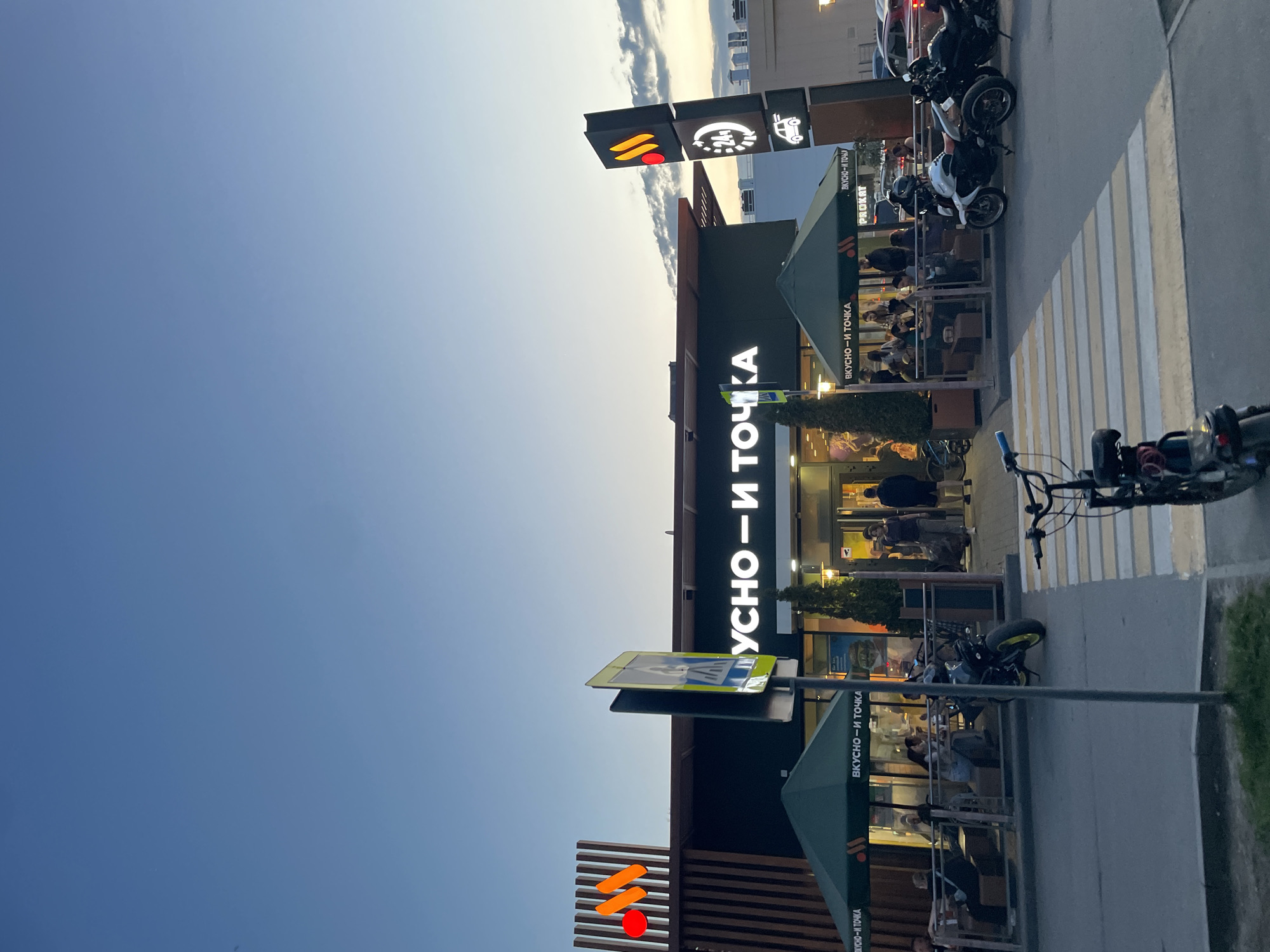 The image shows a restaurant named "Vkusno i Tochka" which translates to "Tasty and That's It" - the Russian successor to McDonald's. The building is modern, with wooden accents and green canopies over outdoor seating areas. There are people dining at the outdoor tables. Parked motorcycles are visible along the sidewalk. Street signs are present. The sky is a gradient of blue to lighter shades near the horizon, indicating either sunrise or sunset. Based on the Cyrillic signage and architecture, the location is likely in Russia, and it is in the city of Kazan.