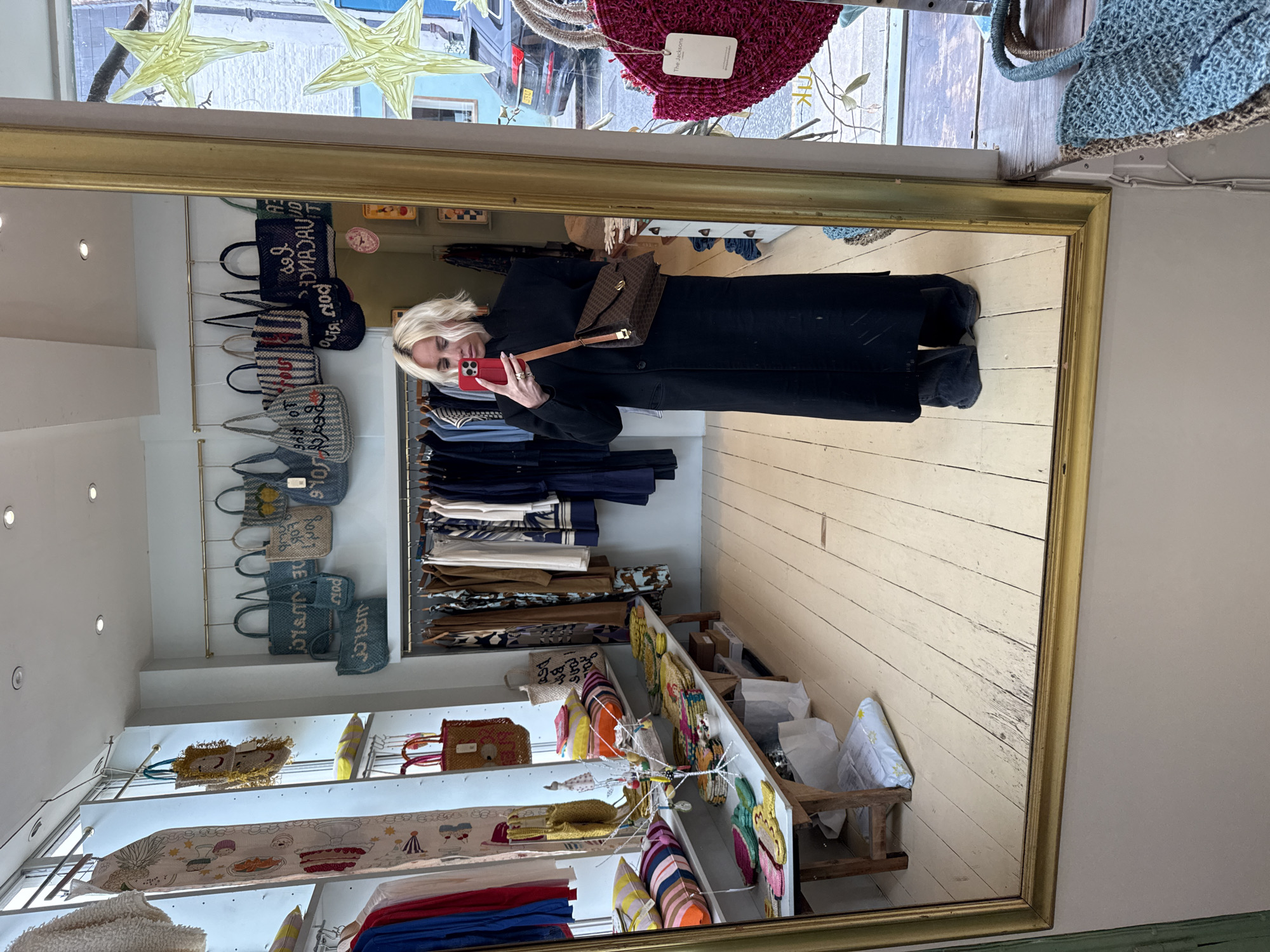 A woman with short blonde hair takes a mirror selfie inside a retail shop located at 231 Portobello Road, London W11 1LT, United Kingdom. She wears a black, high-neck, long coat, holds a red smartphone, and carries a brown patterned shoulder bag with a tan strap. The shop interior features light-colored walls, light wooden floorboards, and recessed ceiling lights. Merchandise displays include clothing racks with various garments, shelves with striped and solid-colored cushions, small woven items, and a metal rod suspending woven tote bags with text such as "LES VACANCES" and "BONJOUR." A red woven bag with a "The Jacksons" tag is visible on the right. The mirror reflects a street scene through the storefront window, showing a dark-colored Audi SUV with a UK license plate, a light blue building facade, and yellow star-shaped decorations hanging in the window.