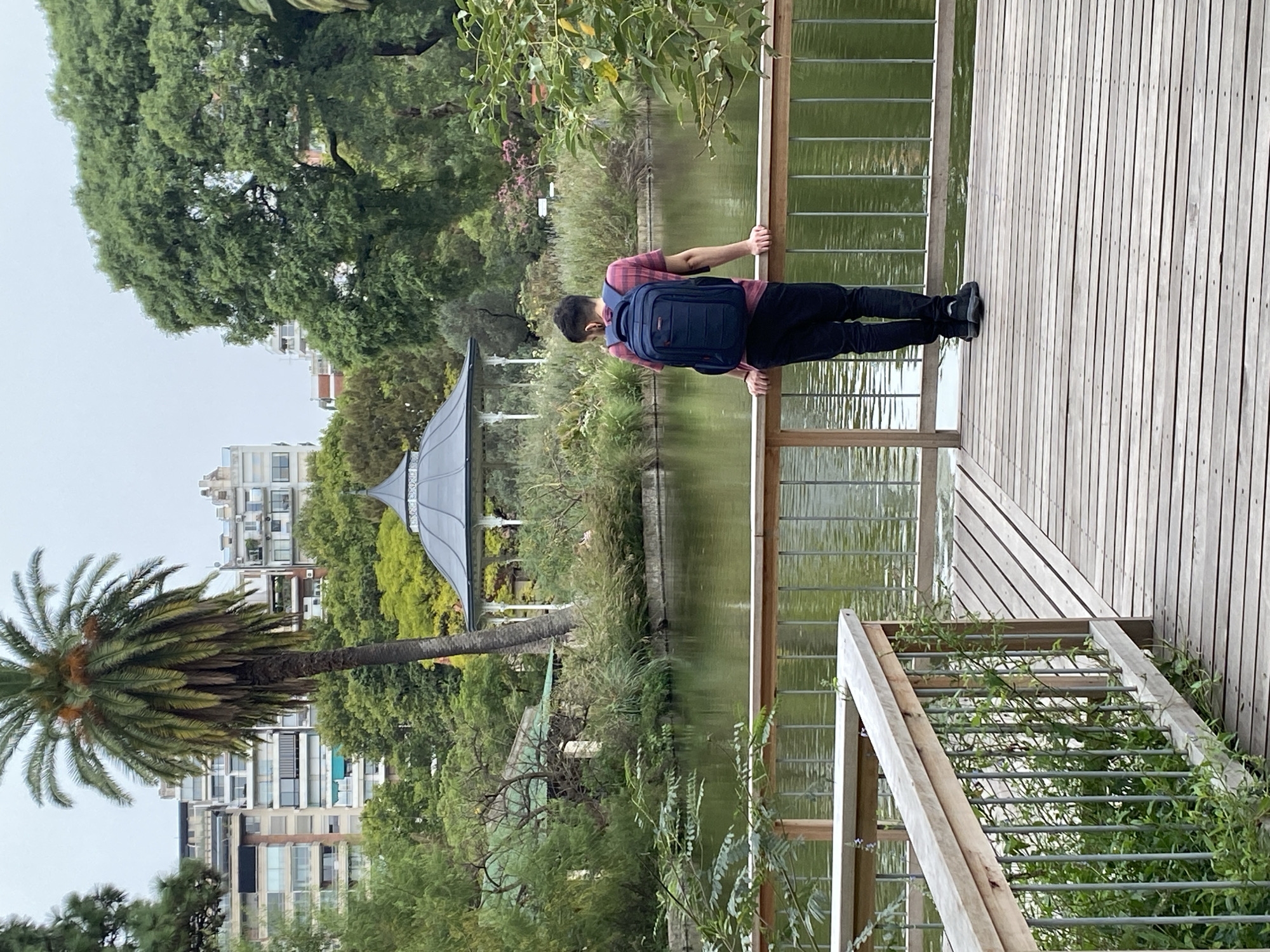 A man with a backpack stands on a wooden deck overlooking a pond in a park in Buenos Aires, Argentina. The pond reflects the surrounding trees and a gazebo. A palm tree is visible on the left side of the frame, and buildings can be seen in the background. The man is wearing a red and black shirt, black pants, and black shoes. The backpack is blue. He is holding onto a wooden railing. Plants grow on the deck and in the pond. The sky is overcast.
