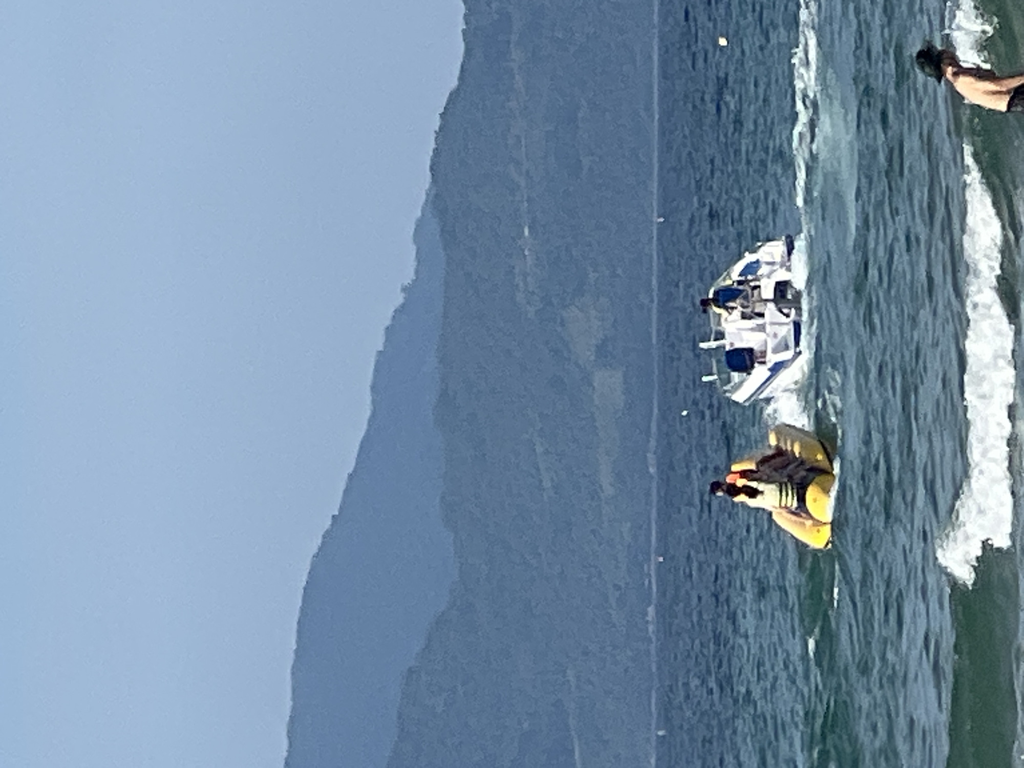 In this image taken from a beach in Đà Nẵng, Vietnam, the ocean is filled with various watercraft and people enjoying a sunny day. In the mid-ground, a yellow banana boat is being pulled by a speed boat, with several individuals visible riding on the banana boat. To the right, another speed boat is cruising through the water, carrying at least one person. In the background, lush green mountains rise behind the expanse of the sea, suggesting a coastal mountainous region. The foreground shows the gentle waves of the ocean breaking on the shore, with a person partially visible on the right edge of the frame, appearing to be interacting with the water. The sky is a clear, light blue, indicating good weather.
