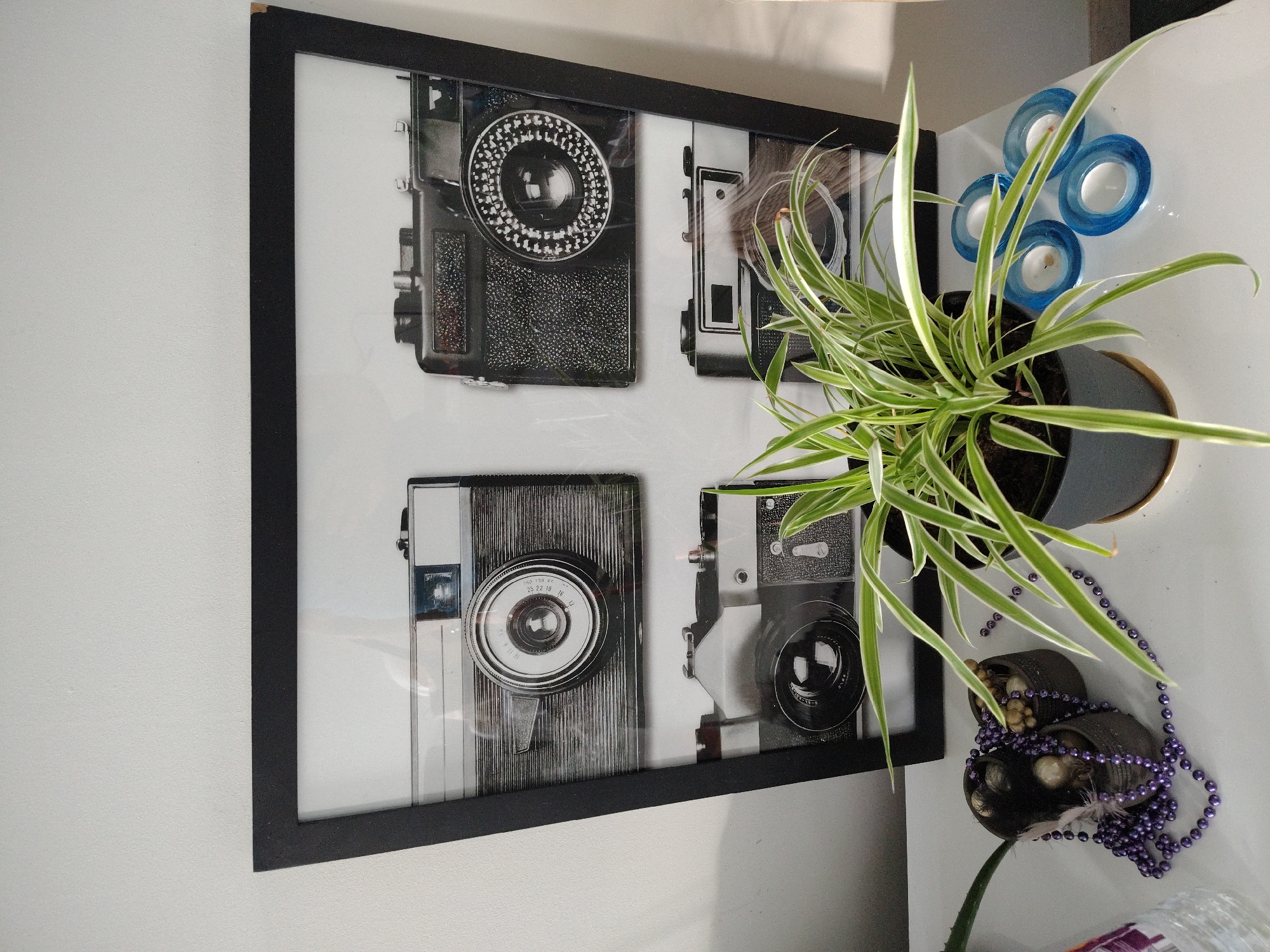This is a vertical, eye-level, medium shot of a framed print of vintage cameras and a spider plant on a white surface. The framed print is a black, trapezoidal frame containing four black and white images of vintage cameras. The top left camera is a silver and black vintage camera. The top right camera is a black vintage camera with a textured body. The bottom left camera is a silver and black vintage camera. The bottom right camera is a black vintage camera. In front of the framed print, a spider plant with green and white striped leaves is in a dark gray pot. To the left of the spider plant, there are two small dark brown pots with a purple beaded necklace draped over them and some small stones inside. To the right of the spider plant, there are four blue, oval-shaped candle holders with small white candles inside. The background is a plain white wall. The scene is likely indoors, possibly in a home or an office.