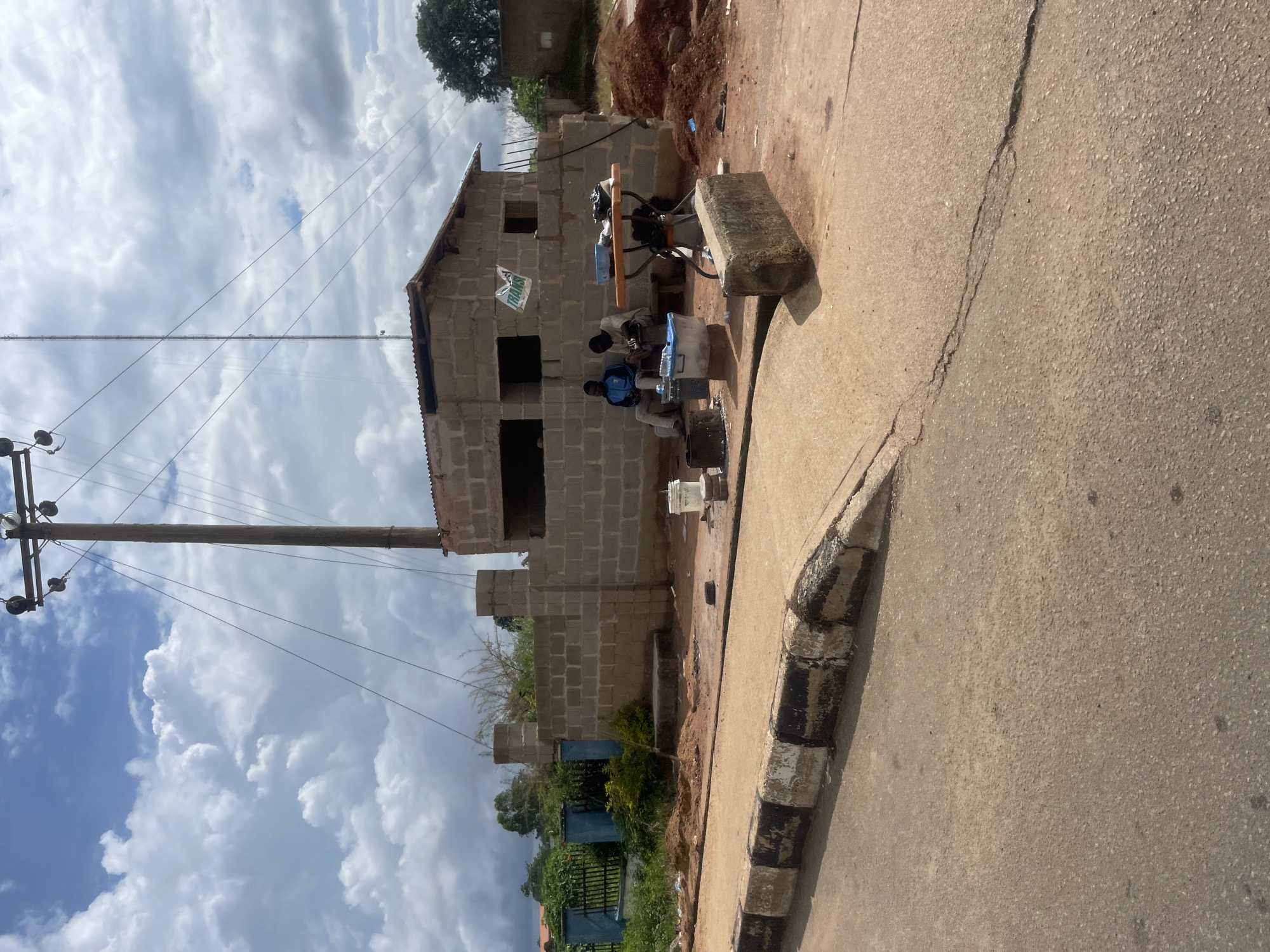 The image depicts an outdoor scene in Bar Jirgi Summa, Nigeria. In the foreground, a paved road with a broken curb runs diagonally. Behind the curb, a partially constructed building made of concrete blocks is visible. The building has an open roof with corrugated metal sheeting. Two men are seated in front of the building, engaged in an activity, possibly a transaction, with items spread out on a table. To the left of the building, a blue fence and lush green vegetation are present. A utility pole with electrical wires stands prominently behind the structure, and a tall antenna-like structure is also visible in the background. The sky is filled with dramatic, puffy white clouds against a blue background. The ground around the building is covered with red soil and some construction debris.