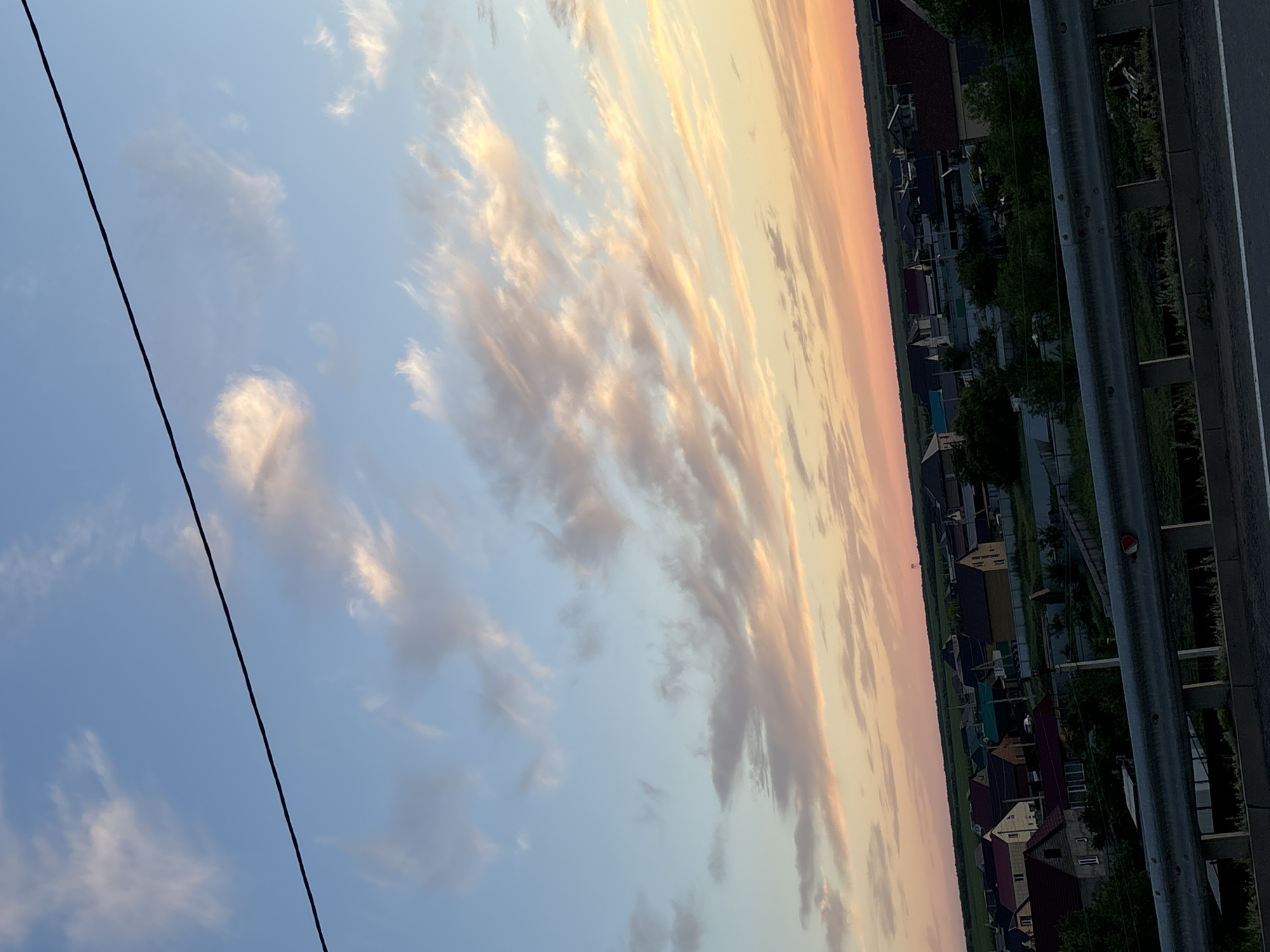 The image shows a village scene at sunset. The sky is filled with clouds in shades of blue, pink, and orange. A power line runs diagonally across the sky. Below the sky is a view of a village with houses and trees. The sun is low on the horizon, casting a warm glow over the scene. The overall mood is peaceful and serene. The image is oriented vertically, and the scene appears to be viewed from an elevated position, possibly a bridge or overpass, as indicated by the presence of a metal railing on the right side of the frame. The houses in the village have pitched roofs and are surrounded by greenery. The time of day is sunset.