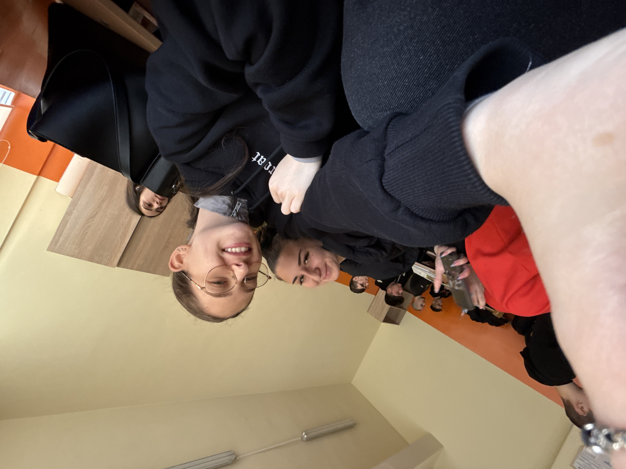This image, originally rotated ninety degrees counter-clockwise, captures an indoor scene of a group of young people, likely in a classroom or common area in Sofia, Bulgaria. The photograph is taken from a low-angle selfie perspective, looking upwards at the gathered individuals.
In the immediate foreground on the right, the light-skinned arm and wrist of the person taking the photo are visible. They wear a black long-sleeved top with ribbed cuffs and a bracelet adorned with dark and light beads.
The central focus of the image is on two young women who are smiling broadly and looking directly towards the camera. The woman on the left, closer to the lens, has light brown hair, wears round, wire-rimmed glasses, and displays a wide smile. She is dressed in a dark hoodie, with a white drawstring visible. On her hoodie, partially obscured white embroidered text is visible, appearing as "t.at" in the original rotated image. To her right, another young woman with darker hair pulled back also smiles towards the camera, wearing a dark top.
Further in the background, several other individuals, predominantly young men, are seated at wooden desks or tables. Most are facing away from the camera, though some can be seen looking in different directions. Their attire is casual and generally dark, with one person wearing a distinctive bright red top. One individual in the far background is seen holding a clear bottle or glass. A black shoulder bag or tote bag rests on a desk in the mid-background.
The setting is characterized by interior walls painted in two distinct tones: a lighter yellow or cream color on the lower section and a vibrant orange on the upper section. Fluorescent light fixtures are visible on the light-colored ceiling. The overall mood appears casual and friendly, with the visible smiles suggesting positive emotions among the subjects interacting with the camera.
