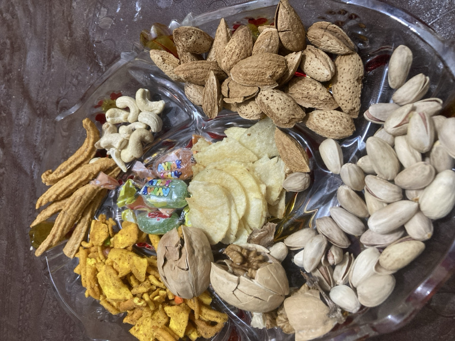 A clear, multi-segmented serving platter, likely glass, is positioned on a dark, patterned surface. The platter contains assorted snack items: a pile of yellow and orange savory snack mix, several golden-brown elongated fried snacks, cashew nuts, almonds in their shells, pistachios in their shells, and plain potato chips. Several individually wrapped, multi-colored hard candies are centrally distributed among the items. Two walnuts are located on the left, one whole and one partially cracked, exposing the kernel. No individuals are visible, and no actions or interactions are depicted. The scene is illuminated, indicating an indoor environment. The image does not contain visual information to confirm the specified location of Quetta City Tehsil, Pakistan.