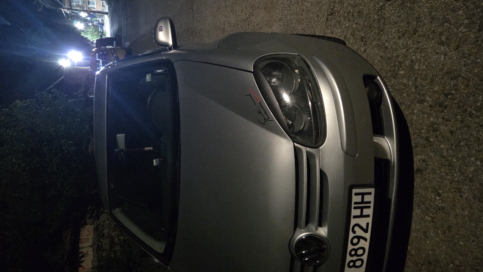 A silver Volkswagen Golf is parked on a street at night in Bania, Bulgaria. The car is facing the viewer at a slight angle, with its front end prominently displayed. A license plate reading "8892 HH" is attached to the front bumper. A decal with script and the words "Sports mind Produced by" is visible on the hood. In the background, streetlights illuminate the scene, and other vehicles are parked further down the street. Trees and buildings line the road, suggesting a residential area. The ground is asphalt.