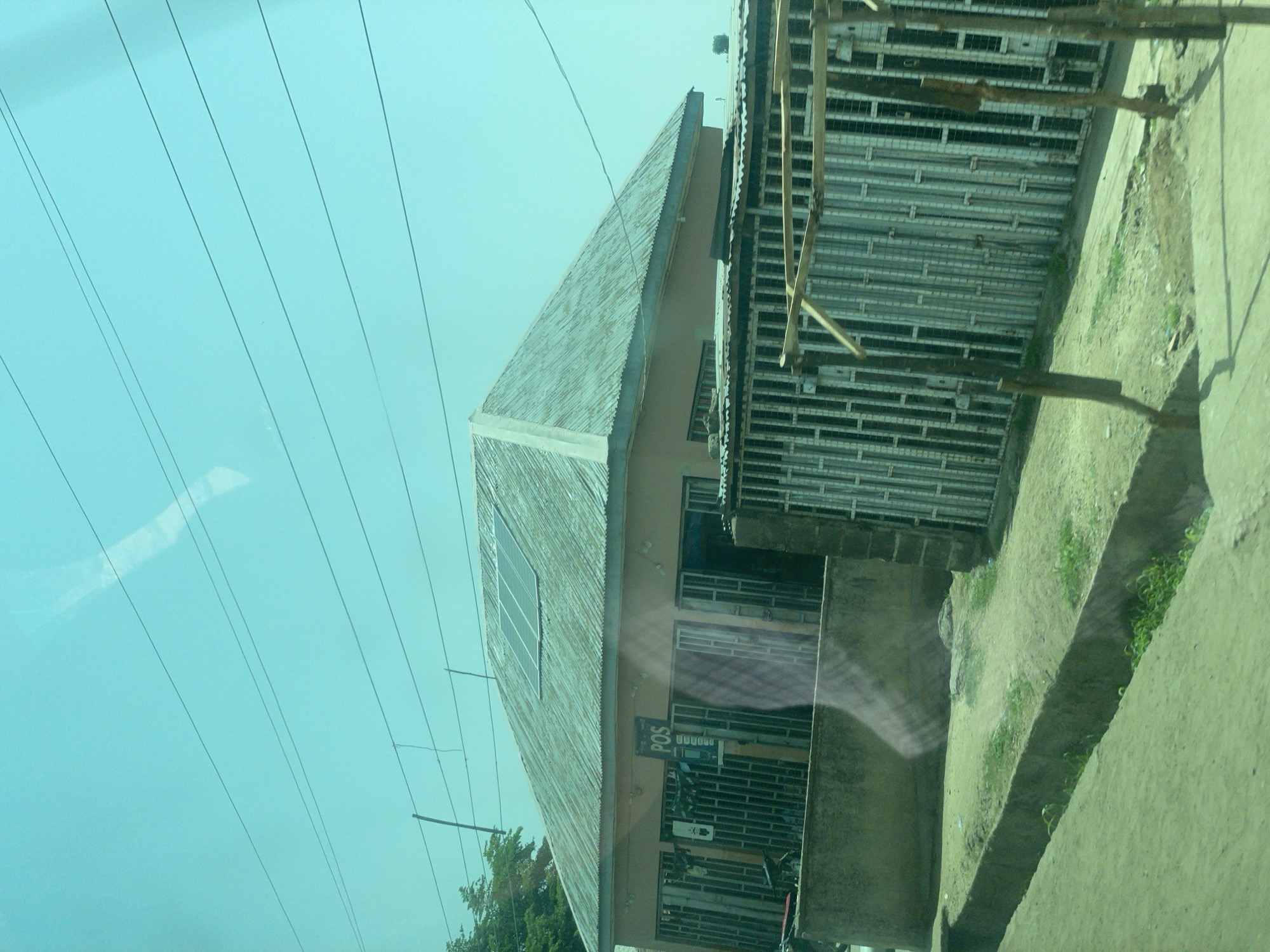 The image depicts a street view in Bauchi, Nigeria, featuring a single-story building with a corrugated metal roof, equipped with solar panels. The building appears to be a commercial establishment, with a prominent "POS" sign visible above its entrance. The storefront is secured with metal grilles, and a bicycle is parked to the left of the entrance. In the foreground, a concrete drainage channel runs along the street, with some vegetation growing within it. The sky is a clear, bright blue, with several power lines crossing overhead. The overall impression is one of a typical urban scene in Nigeria, with a blend of modern technology and traditional architecture.