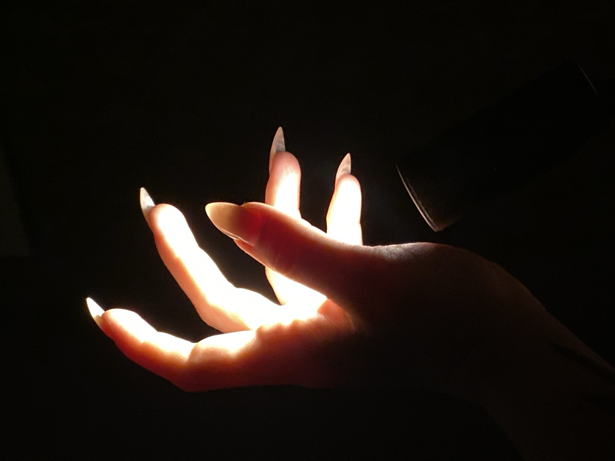 A close-up photograph shows a partially illuminated human hand with long, pointed fingernails against a dark background. The hand appears to be positioned in a reaching or open gesture, with light emanating from between the fingers, creating a stark contrast between the illuminated portions and the deep shadows. There is a dark, indistinct shape in the background, possibly an object partially obscured by the darkness. The environment is unclear, with no identifiable features indicating the location as Chaikovsky, Russia, or any other specific place. No people are visible other than the hand itself.