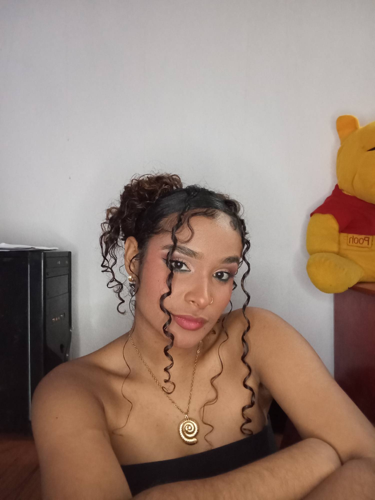 The image depicts a female subject situated indoors in Juan Griego, Venezuela. The subject has light brown skin and dark, curly hair styled partially up, with several ringlets framing her face and resting on her shoulders and chest. She is wearing visible eye makeup, including eyeliner and eyeshadow, and light pink lip color. A small gold stud earring is present in her right ear, and a small gold nose ring adorns her right nostril. She wears a gold chain necklace with a large, spiral-shaped pendant. Her shoulders are bare, suggesting a strapless top or dress of a dark color. The subject gazes directly forward with a neutral expression. In the background to the left, a black computer tower is visible, with white papers or objects stacked on its top. To the right, a yellow Winnie the Pooh plush toy, wearing a red shirt with the word "Pooh" (inverted), is partially visible, positioned on a dark red-brown surface. The wall behind the subject is plain white.