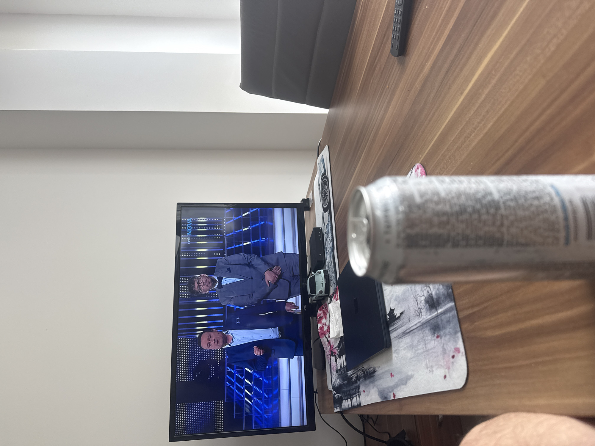A television screen shows two men in suits against a blue, geometric patterned background. A can is in the foreground, partially obscuring the view of the television. The can is resting on a wooden surface, next to a mousepad with a landscape design. Other objects on the surface include a remote control and a computer. The location is Sofia, Bulgaria.