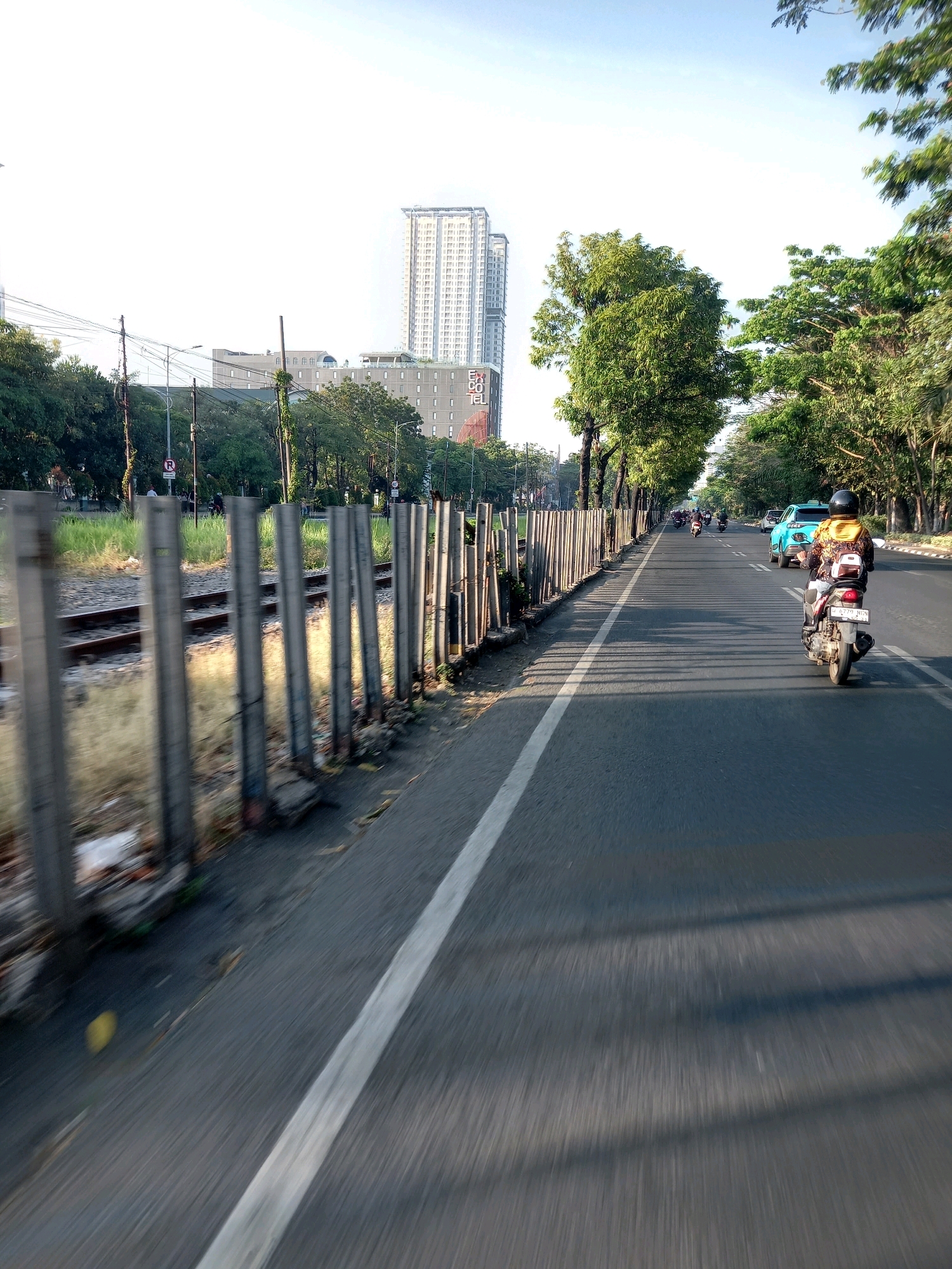 The image depicts a roadside scene in Surabaya, Indonesia, likely during the daytime given the bright sunlight and shadows. A paved road stretches into the distance, with a motorcycle in the foreground and a turquoise car further ahead. The road is bordered by a fence made of vertical posts and grassy areas, with railroad tracks visible behind the fence. Trees line the right side of the road, and buildings, including a tall skyscraper, are visible in the background. The overall impression is a busy urban environment with traffic and infrastructure.