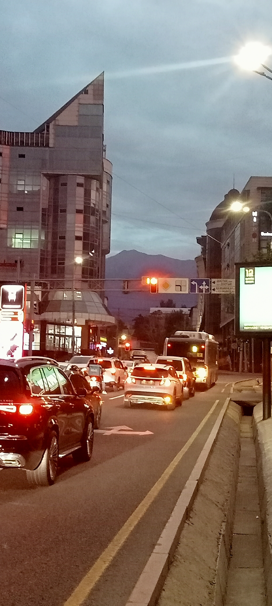 The image captures a street scene in Almaty, Kazakhstan, during what appears to be either dawn or dusk, with a traffic light displaying a red signal. A significant traffic jam is evident, with multiple cars and a bus stationary on the road. In the foreground, a dark-colored sedan is visible from the rear, its brake lights illuminated. Further down the street, other vehicles, including a white SUV and a bus, are also stopped.
The cityscape features modern architecture, notably a distinctive angular building with large glass windows on the left side of the frame. In the background, a mountain range is faintly visible against a cloudy sky, a characteristic geographical feature of Almaty. The street is paved, with clear lane markings and a concrete drainage ditch along the right side. Streetlights and building lights are on, suggesting low ambient light conditions. The overall atmosphere is one of urban congestion and impending movement as the traffic light is red.