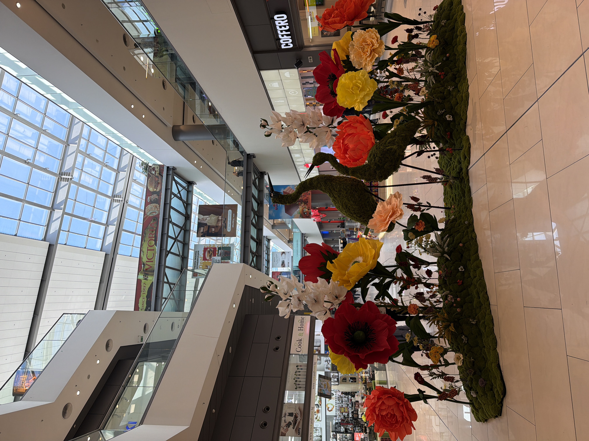 Captured from an elevated perspective, the image depicts the interior of a shopping mall, notably featuring a sprawling floral arrangement. This arrangement is comprised of large artificial flowers in shades of red, orange, and yellow, interspersed with mossy bird-shaped topiaries, all set against a verdant base of moss.
The mall's architecture is modern with multi-level floors connected by escalators, all bathed in natural light filtering through a large glass ceiling. Retail storefronts are visible in the background, bearing brand names like "COFFERO," "Cook & Home," and "GEFU". Banners hanging from the ceiling promote "NEW VICTORIA" and "Deal". The image appears to be taken inside a shopping mall in Sofia, Bulgaria, due to visible Bulgarian text on retail signs and banners. The overall ambiance is bright, spacious, and suggestive of a commercial environment.