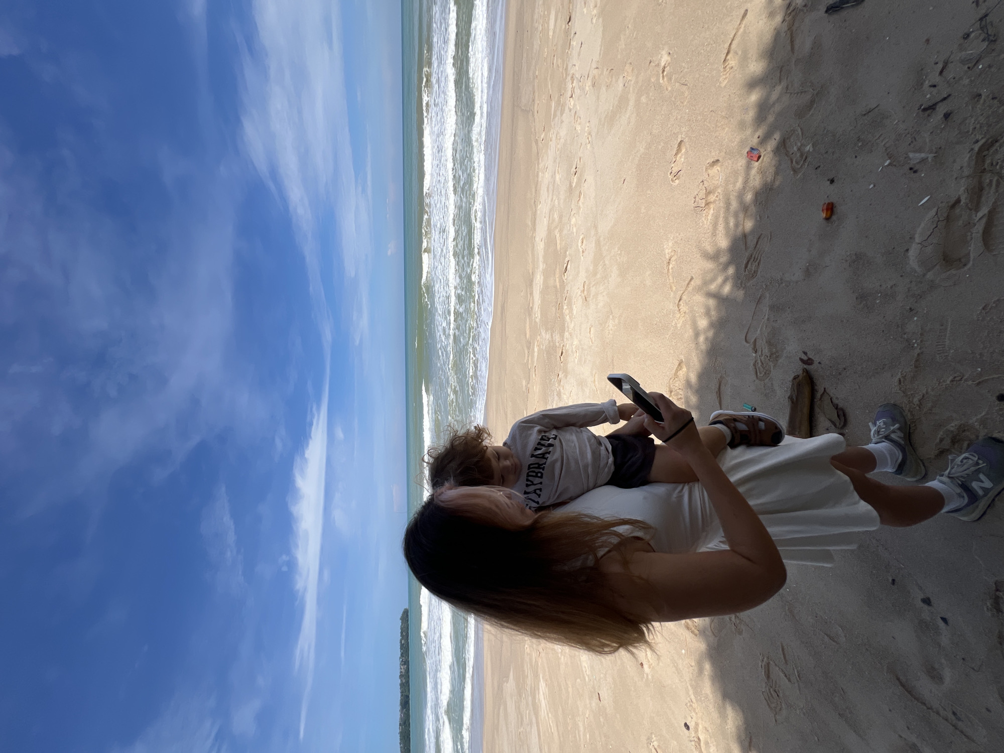 A woman stands on a sandy beach, holding a young child. She is wearing a white dress and sneakers, and the child is in a grey shirt and dark shorts. Both are looking at a smartphone in the woman's hand. The ocean with gentle waves is in the background, under a bright blue sky with scattered clouds. The scene is outdoors, likely a coastal area.