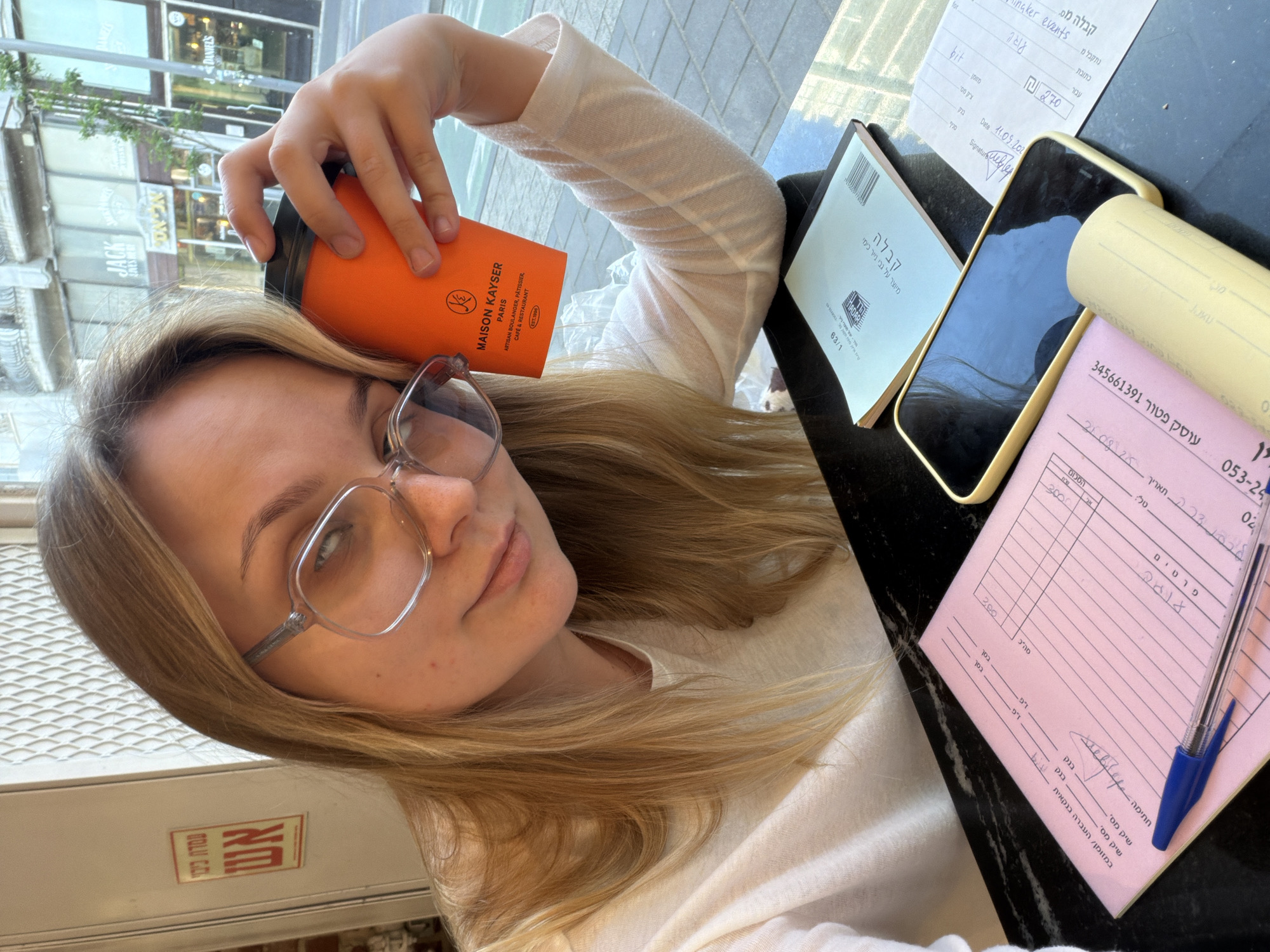A young, fair-skinned woman with long blonde hair and glasses is posing for a selfie while holding an orange Maison Kayser coffee cup. The Maison Kayser logo is clearly visible on the cup. Several items are on the table in front of her, including a pink receipt with Hebrew text, a blue ballpoint pen, a smartphone, a yellow paper roll, and a notebook with the word "Kavala" in Hebrew printed on the cover. There is also a restaurant or cafe receipt with the date "11.09.202" written on it, along with a price of 270 NIS. The background reveals the reflection of a street with storefronts, one of which is "Jack Daniel's." A red sign in Hebrew is also visible. The location appears to be a cafe in Tel-Aviv, Israel, judging by the Hebrew writing and overall ambiance. The woman is likely a customer at the cafe.