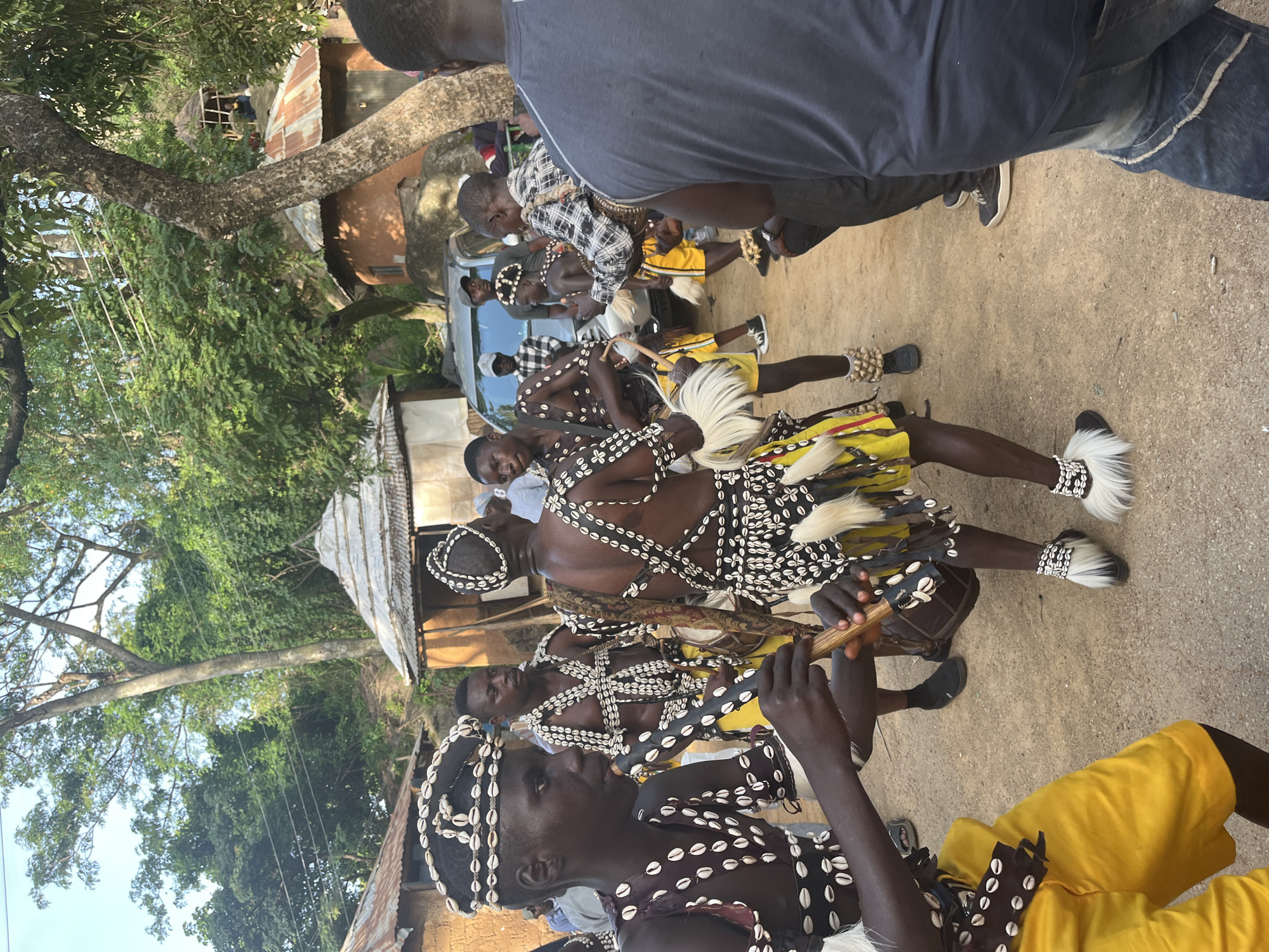 The image captures a group of young men in traditional attire, adorned with shells and white fur accents, participating in a cultural performance or ritual. The central figures are dressed in elaborate costumes, including headdresses, vests, and skirts, with intricate shell embellishments. One individual in the foreground is playing a wooden instrument, possibly a flute or a rattle, while others appear to be dancing or drumming. Several of the performers are wearing white fur leg coverings and anklets. In the background, traditional round huts with thatched roofs are visible, characteristic of rural settlements in areas like Jos, Nigeria. A silver SUV is parked behind the performers, suggesting a blend of traditional and modern elements within the community. The environment is dusty and appears to be outdoors, with trees and foliage indicating a warm climate. The overall scene depicts a lively cultural gathering.