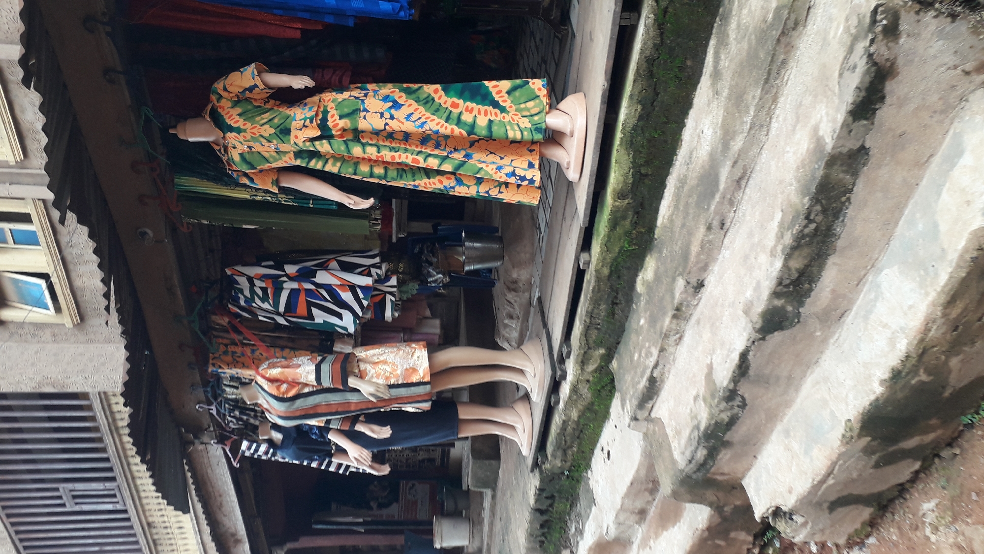 This is a medium shot, eye-level, outdoor photograph of a street-level shop in Nkpor, Nigeria. The shop has a covered awning area where clothes are displayed on mannequins. In the foreground, three mannequins are visible. The mannequin on the far left wears a dark blue garment with white stripes and a black skirt. The middle mannequin is dressed in a patterned orange and striped dress. The mannequin on the right displays a long, green and orange patterned dress. Behind the mannequins, various clothing items hang from racks under the awning. A multi-story building with barred windows is visible above the shop. The ground in front of the shop is made of concrete steps, some of which are covered in moss. The overall scene depicts a retail environment in an urban setting.