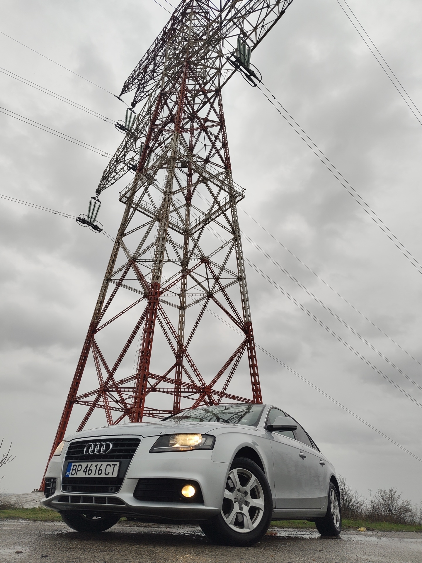 A low-angle shot captures a silver Audi A4 (B8 generation) parked on a wet asphalt surface. The vehicle's front left quarter is visible, with its headlights and front right fog light illuminated. The Bulgarian license plate "BP 4616 CT" is prominently displayed on the front bumper.
Dominating the background is a massive, multi-sectioned lattice steel electricity transmission pylon, painted in alternating reddish-brown and white sections. This pylon extends high into a uniformly overcast, gray sky. Multiple high-voltage power lines, suspended by green and white ceramic insulators, originate from the pylon and traverse horizontally across the frame.
The ground around the car is visibly damp, suggesting recent precipitation. Sparse, dark-toned vegetation is visible on the slightly elevated ground to the right of the vehicle. No individuals or specific actions are depicted. The license plate's "BP" prefix indicates registration in Vratsa province, Bulgaria. Given the presence of a large-scale power transmission pylon in Vratsa province, the scene is consistent with an outdoor location in the Kozloduy area of Bulgaria, known for its nuclear power plant and associated high-voltage infrastructure.