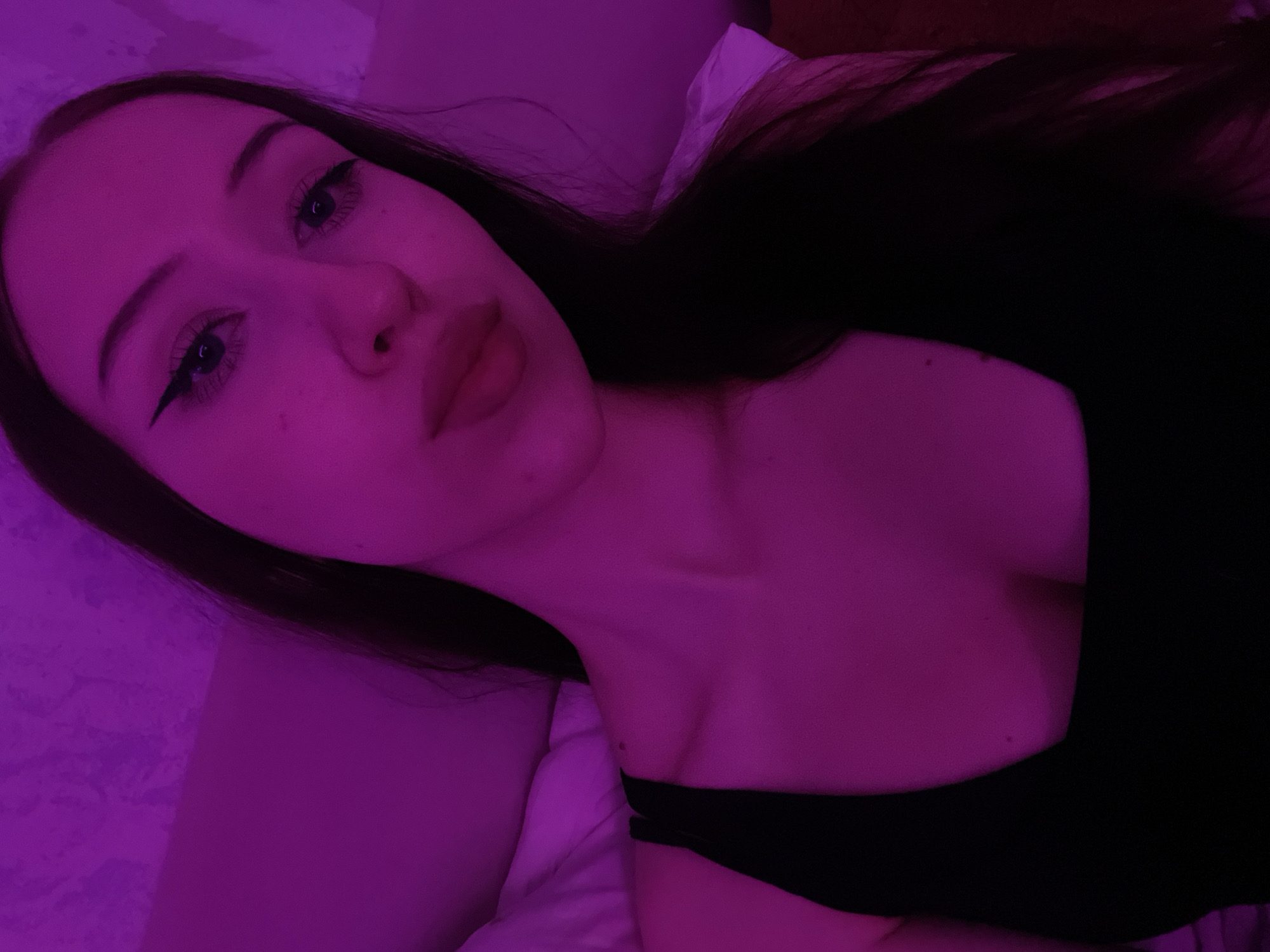 A young woman with dark hair and a black top is posing for a selfie. She is illuminated by purple light. The background appears to be a textured wall, possibly in a bedroom. The woman has eyeliner and mascara on, and her lips are parted slightly. She is looking directly at the camera. There is no indication of the location being Votkinsk, Russia.