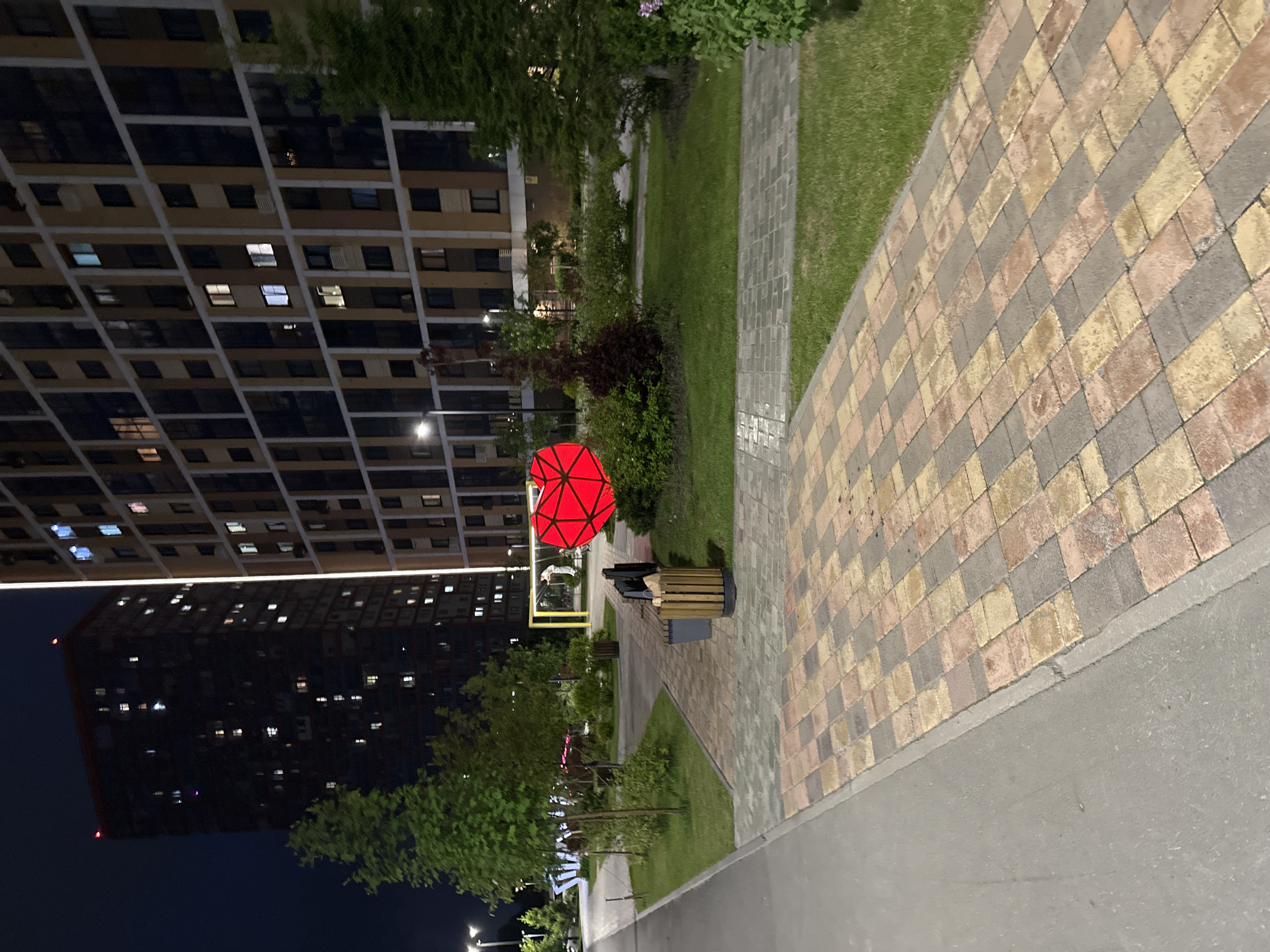 The image shows a view along a pathway in an urban setting at night. On the left side, there are several tall, dark buildings with many windows, some of which are lit. Adjacent to the pathway is a strip of grass and greenery, including trees and bushes. A red, geometric sculpture, possibly a heart, stands prominently. A wooden and metal bench is nearby. The pathway is paved with patterned bricks, and a concrete sidewalk runs along the right edge of the image. Further down the path, lights and more greenery are visible. The sky is dark, indicating it is nighttime.