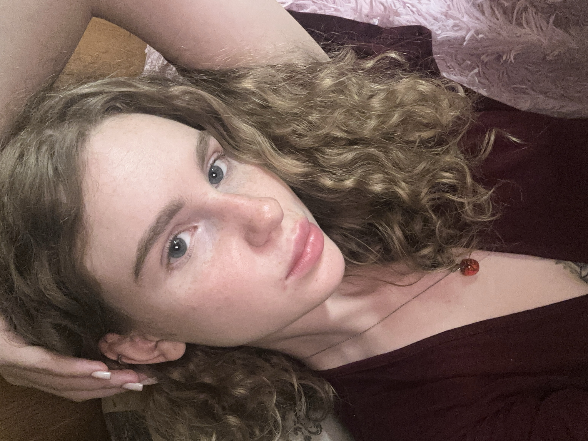 A close-up, angled portrait of a fair-skinned individual with curly blonde-brown hair and light blue eyes. The person is lying down, with their head resting on their hand. The hand has manicured nails with a neutral polish. Freckles are visible on their face. They are wearing a dark red top and a necklace with a red bead. A pink, fluffy blanket is visible in the background, partially obscured by their hair. No location information is discernable from the image.