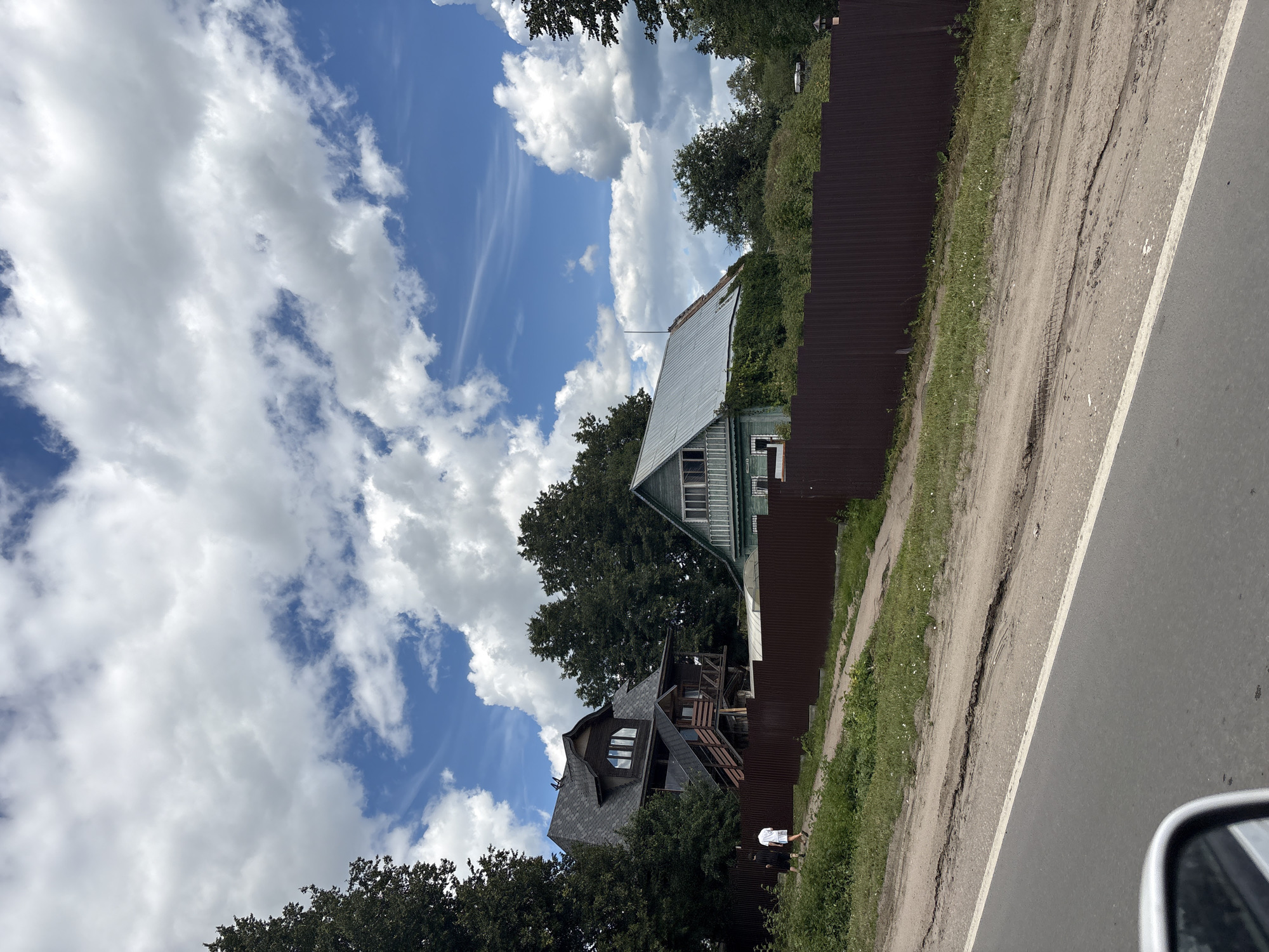This image, taken in Pereslavl-Zalessky, Russia, depicts a scene from a car's perspective, showing a residential area by a road on a cloudy day. The road is paved with a white line marking the edge, and to its left is a dirt path with grass. A tall, dark brown corrugated metal fence runs along the path, separating it from the properties behind.
Behind the fence are two houses. The house closer to the foreground is dark with a complex, multi-gabled roof covered in dark shingles. It has a wooden, multi-level balcony structure attached. Further back and slightly to the right is a lighter-colored house with a light-colored metal roof and green siding. Both houses are surrounded by dense green trees.
In the distance, the sky is a vibrant blue filled with dramatic, fluffy white clouds. The overall impression is of a rural or suburban setting, possibly a dacha village, under a dynamic sky. The image is oriented vertically, making it appear as if the viewer is looking up or at a tilted perspective. The time of day appears to be daytime, given the bright sky and the way the light falls on the scene. There is no visible text in the image. A person in a white t-shirt and dark shorts is visible walking along the dirt path.