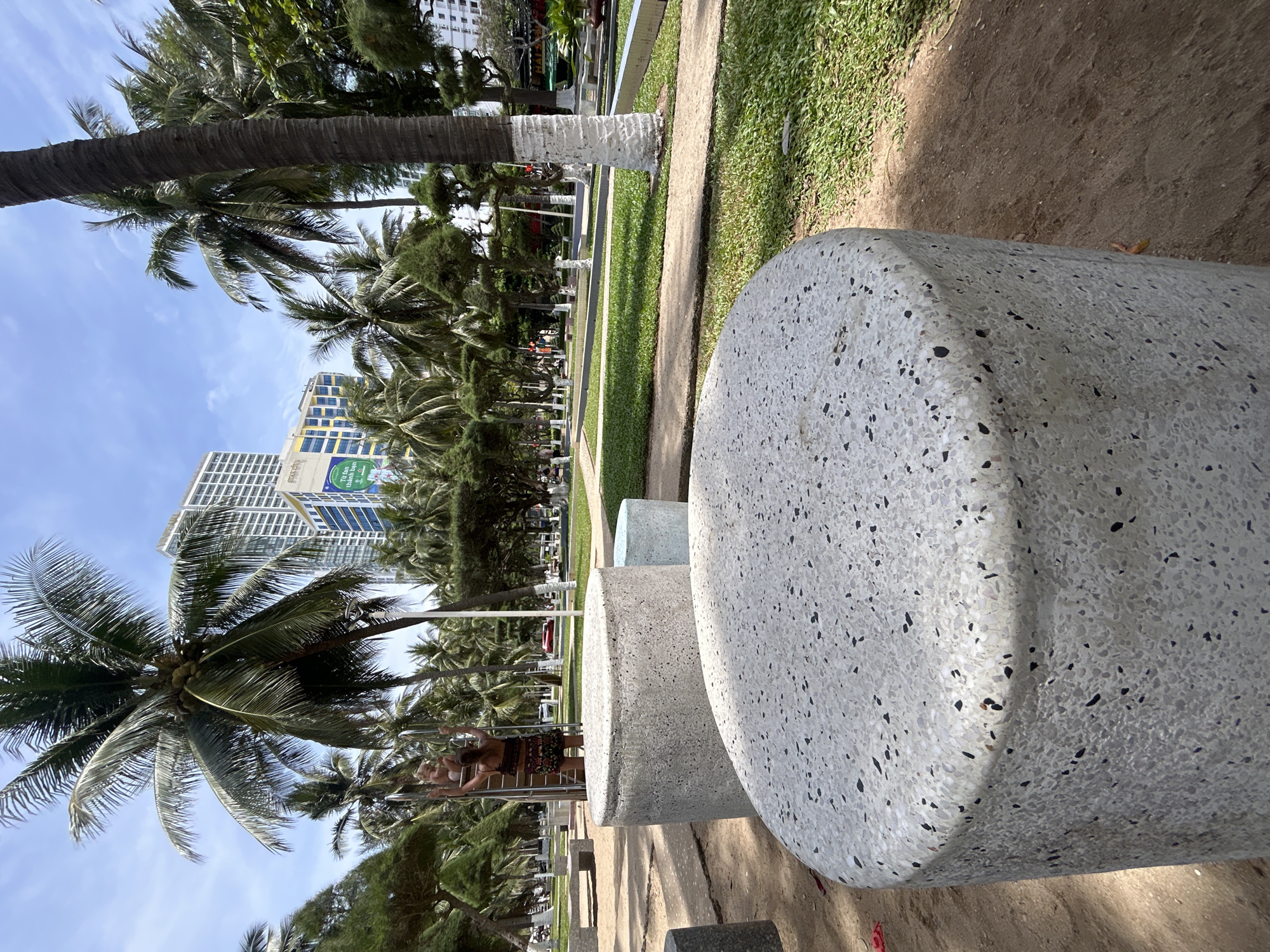 The scene is set in Nha Trang Ward, Vietnam, featuring a seaside park with palm trees and modern architectural elements. In the foreground, there are cylindrical stone benches with a speckled pattern. A woman wearing a skirt and top is seen near the benches, with other people visible further down the path. The park includes a grassy strip, a walkway, and trees that line the path. In the background, modern buildings rise against a blue sky.