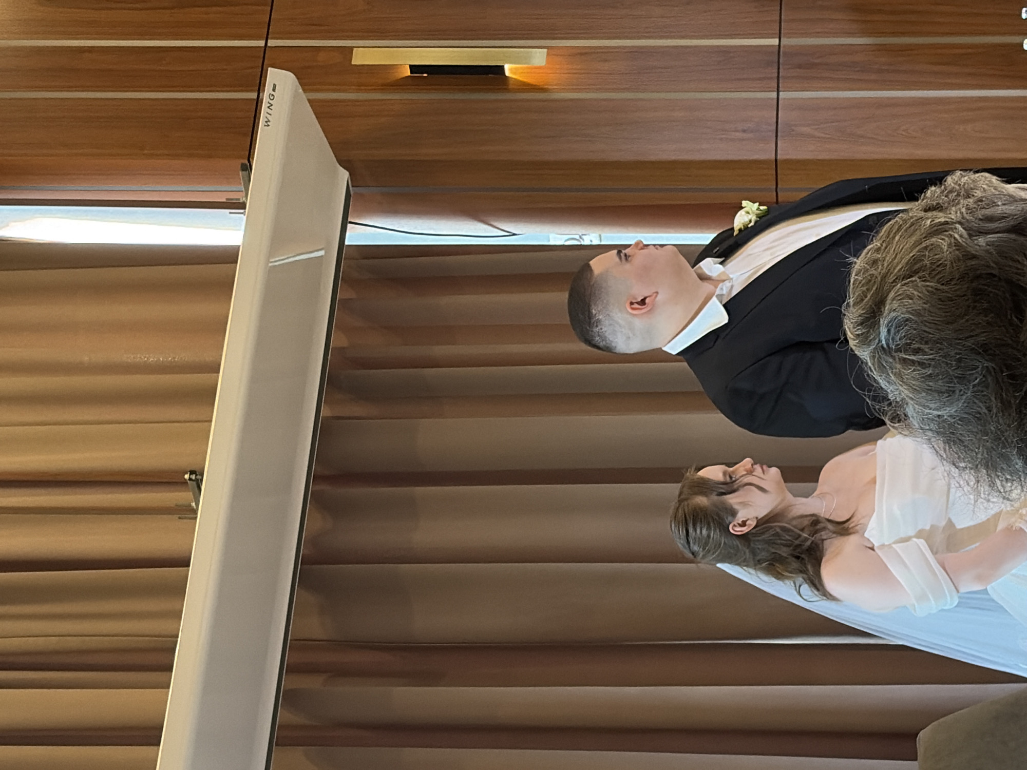 A wedding scene, likely in an indoor venue in Yekaterinburg, Russia, shows a bride and groom. The groom is wearing a black suit and the bride is in a white wedding dress. They are standing closely together. The background features wooden paneling and a vertically mounted, white projector screen marked "WING."