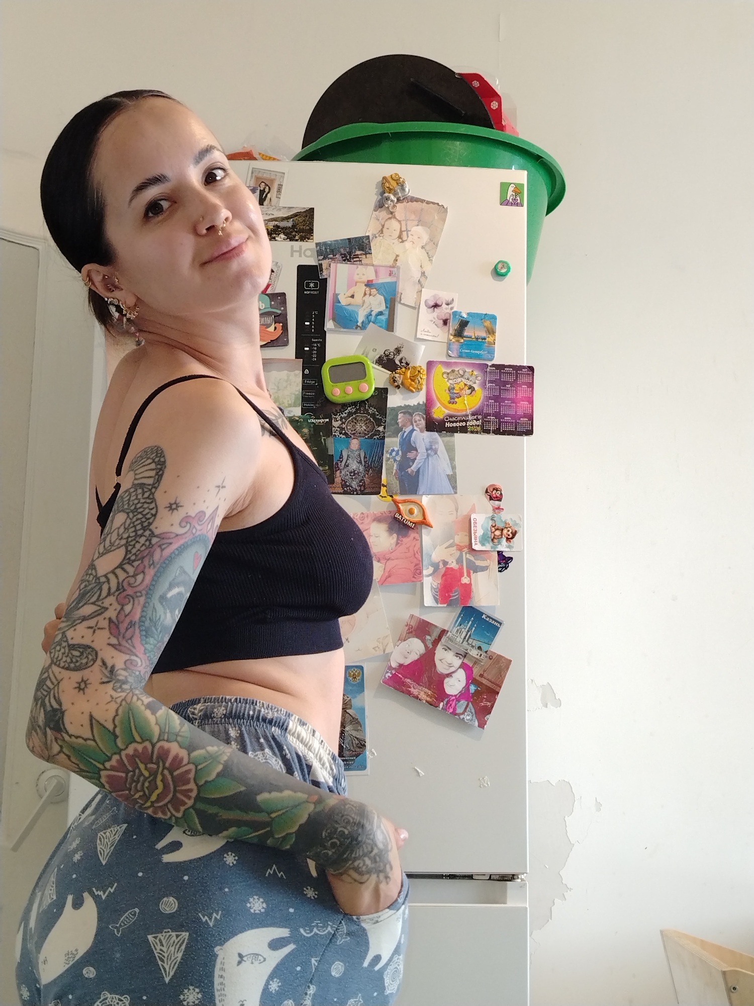 A female subject is depicted indoors, positioned in front of a white refrigerator in the city of Noviy, Russia. The subject, with dark hair pulled back and multiple ear and nose piercings, wears a black ribbed crop top and blue pajama bottoms patterned with white bears, fish, and snowflakes. Her arms are extensively tattooed with designs including a snake-like pattern, stars, and a prominent floral motif on her left forearm. She looks toward the camera with a slight smile, her left hand partially obscured near her hip. The white refrigerator behind her is adorned with numerous magnets and photographs, including images of children, a wedding couple, calendars, a green digital timer, and magnets referencing Saint Petersburg, Batumi, and Kazan. A visible panel on the refrigerator displays "NO FROST," "-16°C," and "-18°C." A green plastic basin containing a black and red object rests on top of the refrigerator. The white wall adjacent to the refrigerator exhibits areas of peeling paint. A light-colored wooden object is partially visible in the bottom right corner of the frame.