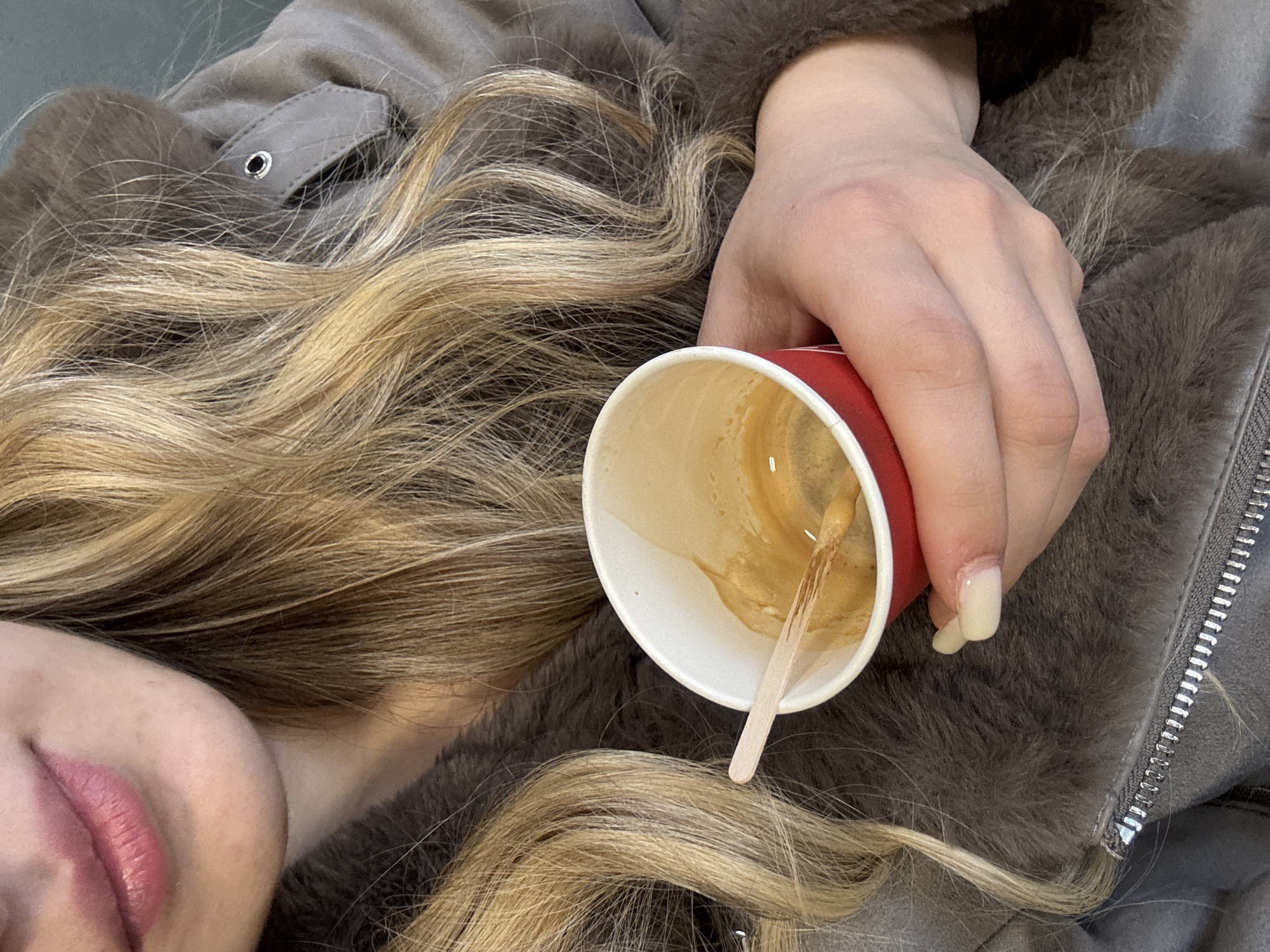 A photograph shows a person lying down, possibly indoors, holding a red coffee cup with a wooden stirrer inside. The cup contains the remnants of a coffee beverage. The person has blonde, wavy hair spread across a gray jacket with fur trim. A small portion of the person's face, specifically the lips, is visible. A zipper is visible near the right edge of the jacket. The precise location within Yambol, Bulgaria is not discernable from the available information.