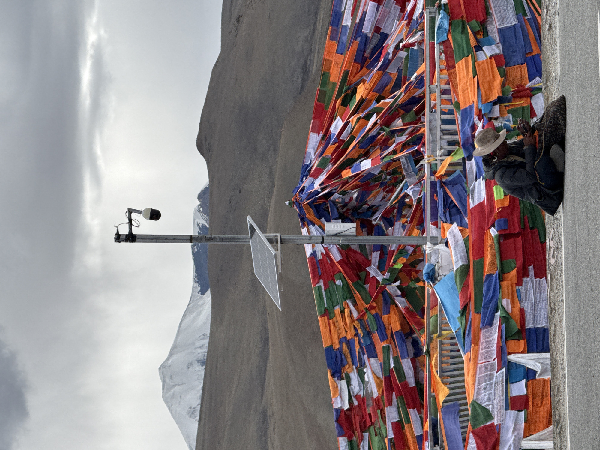 The image captures a vibrant, high-altitude scene in Lhatsé, China, characterized by colorful Tibetan prayer flags and a rugged mountainous backdrop. The original image is oriented on its side, but when viewed correctly, the prayer flags dominate the right and central portions of the frame.
Numerous rectangular prayer flags, primarily in blue, white, red, green, and yellow, are tightly strung together, creating a dense, multicolored tapestry that billows slightly. Many of the white flags feature visible black text, indicative of Tibetan mantras or prayers. To the far right, partially obscured by these flags and a concrete pillar, a person is visible. They wear a light-colored straw hat, a dark jacket, and are draped in a patterned blanket or shawl. The individual appears to be holding a device, possibly a camera or smartphone, with their hands raised, suggesting they are observing or photographing the scene.
On the left side of the frame, a metal pole stands, equipped with a security camera or monitoring device at its apex and a small, tilted solar panel mounted beneath it, indicating modern infrastructure in this remote location. Behind these elements, imposing mountains rise under an overcast sky. Several peaks are distinctly snow-capped, suggesting cold temperatures and high elevation. The sky is a uniform gray, broken by streaks of lighter cloud formations. The overall impression is one of cultural tradition and natural grandeur coexisting with technological elements.