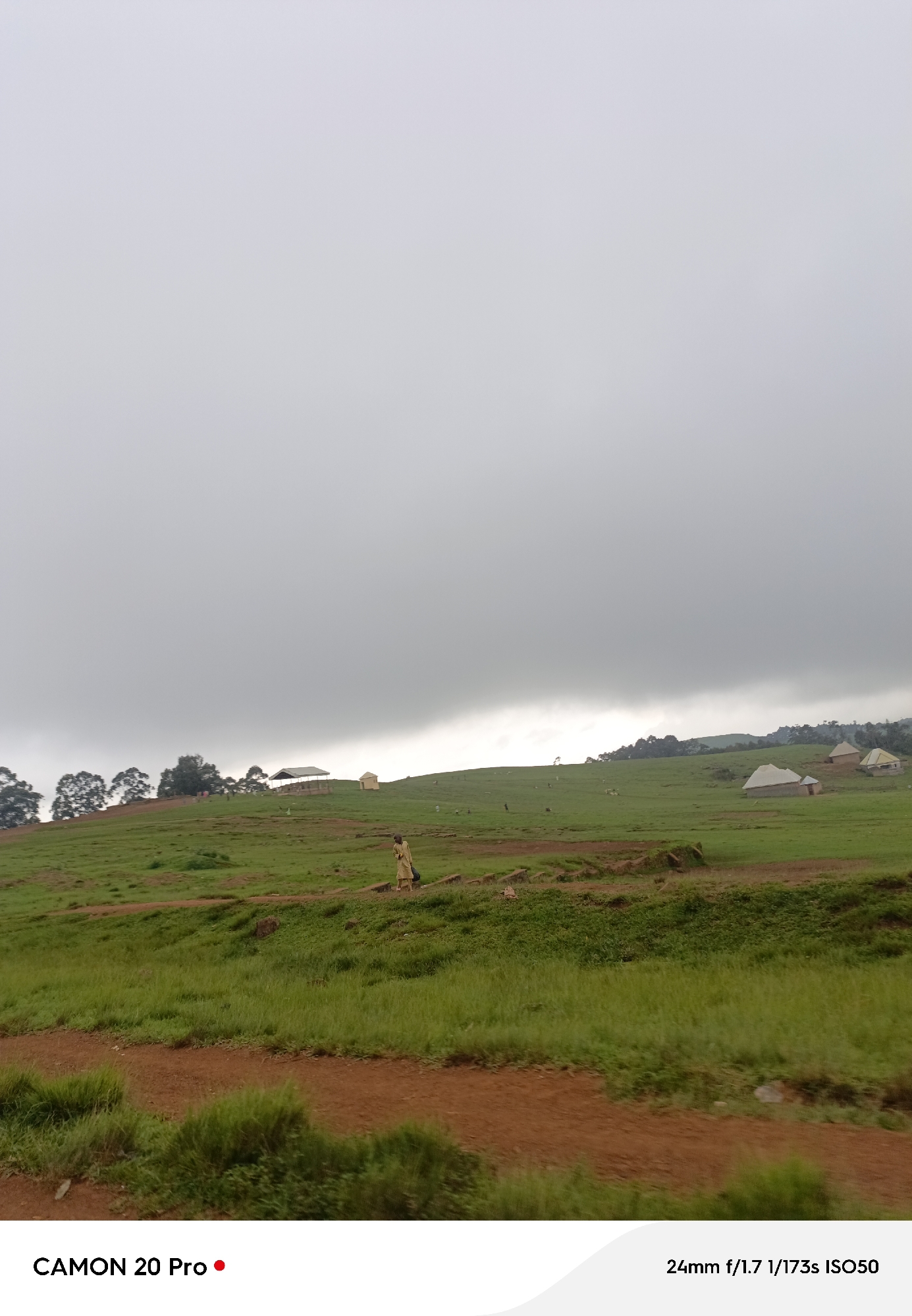 The image shows a rural landscape under a heavily overcast sky. In the mid-ground, a person wearing a light-colored garment walks on a dirt path that winds up a grassy hill. Several small structures, possibly huts or shelters, are visible scattered across the rolling green hills. In the far distance, a line of trees forms the horizon against the grey sky. The foreground consists of a rough, reddish-brown dirt track with patches of green grass. The text overlay in the bottom left corner reads "CAMON 20 Pro" with a red dot, and in the bottom right corner, it displays camera settings: "24mm f/1.7 1/173s ISO50". The scene appears to be a tranquil, everyday moment in a village or farming community in Nguroje, Nigeria.