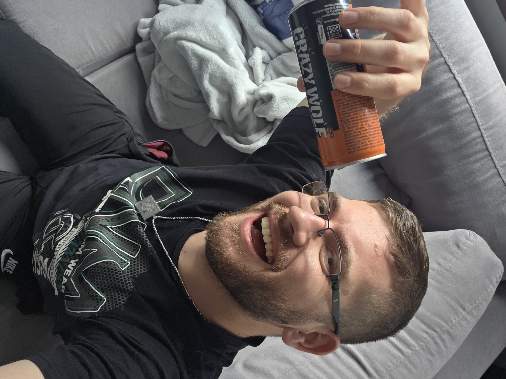 A man is lying on a grey couch, holding an orange and black can of "Crazy Wolf" energy drink. He is wearing glasses, a black t-shirt with a grey and green design, and black pants. He has a beard and is smiling broadly, showing his teeth. A Nike logo is visible on his shirt sleeve. A white blanket is behind him on the couch. The location cannot be determined as Yagodovo, Bulgaria from provided images.